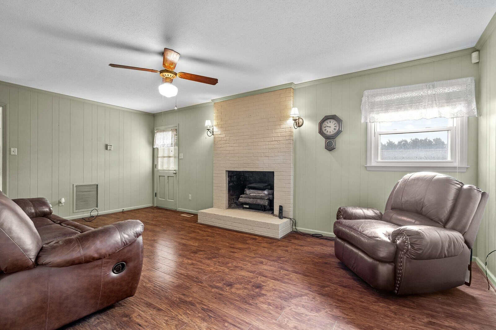 Living room interior with hardwood floors, pastel green walls, two leather armchairs, a fireplace, and a ceiling fan.