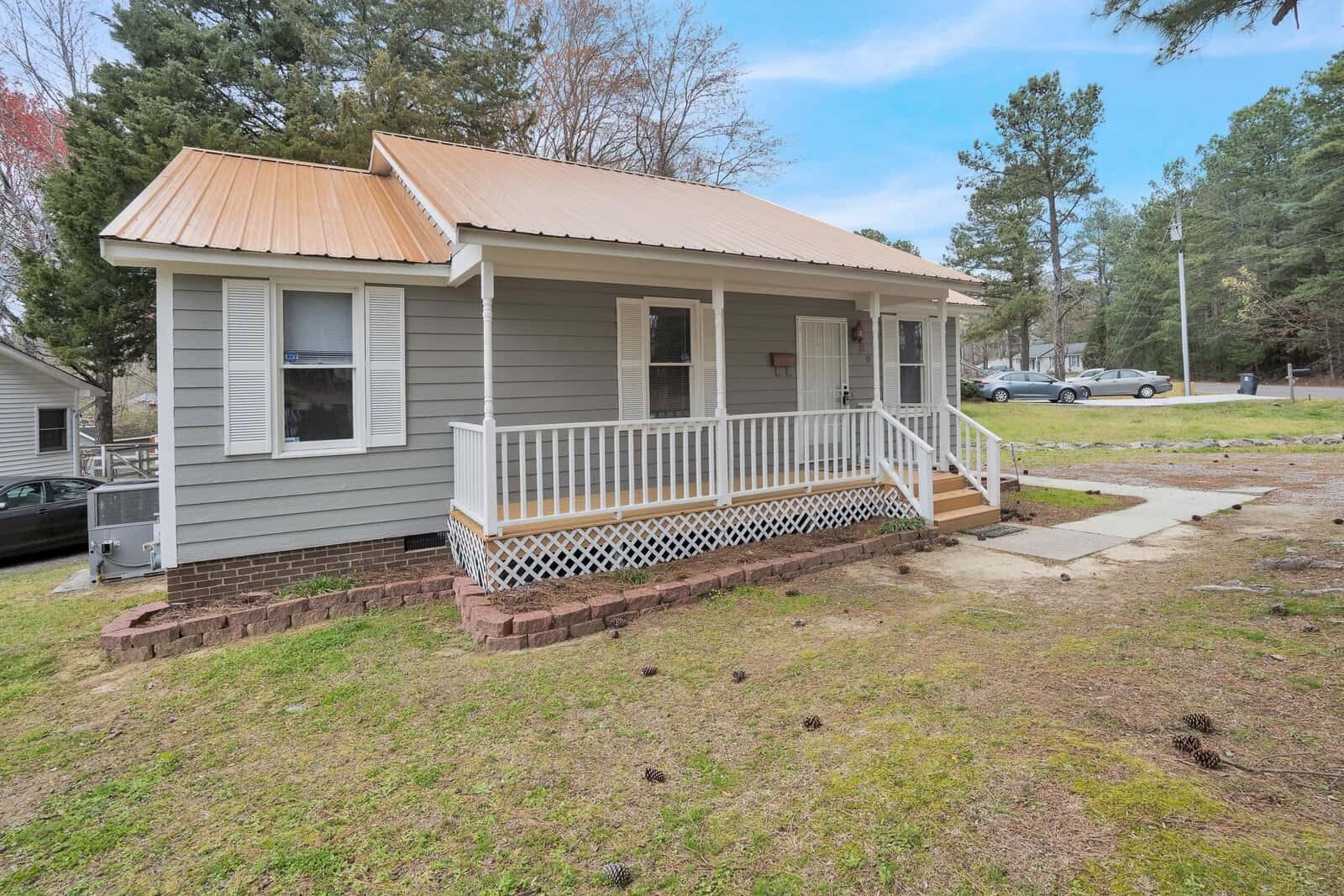 A small single-story house with a white exterior, metal roof, and a front porch with wooden railings, situated on a lot with patchy grass. there is a concrete pathway leading to the porch and some cars visible in the background to the left.