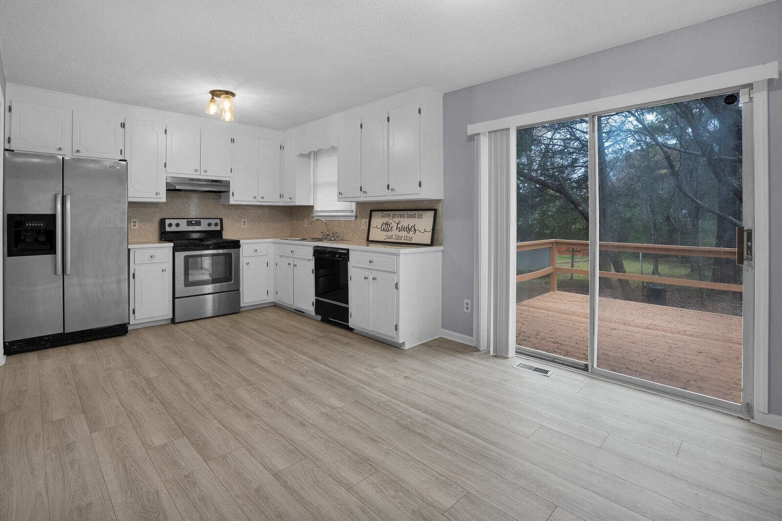 Modern kitchen with stainless steel appliances and white cabinets, featuring a sliding glass door that opens to a deck with a view of trees.