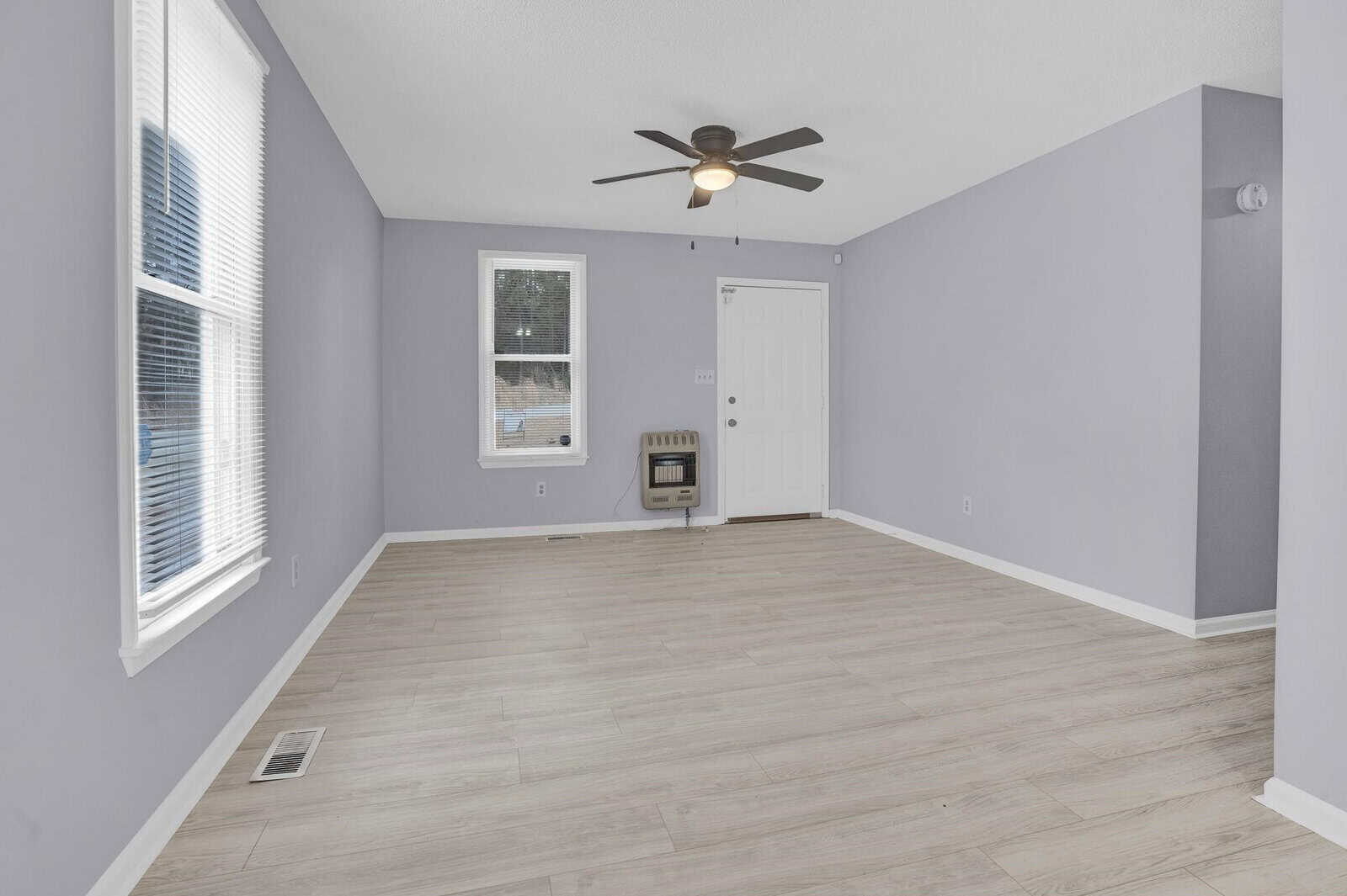 An empty room with gray walls, laminate flooring, and a ceiling fan. a heater is placed near the doorway, with two windows allowing natural light into the space.