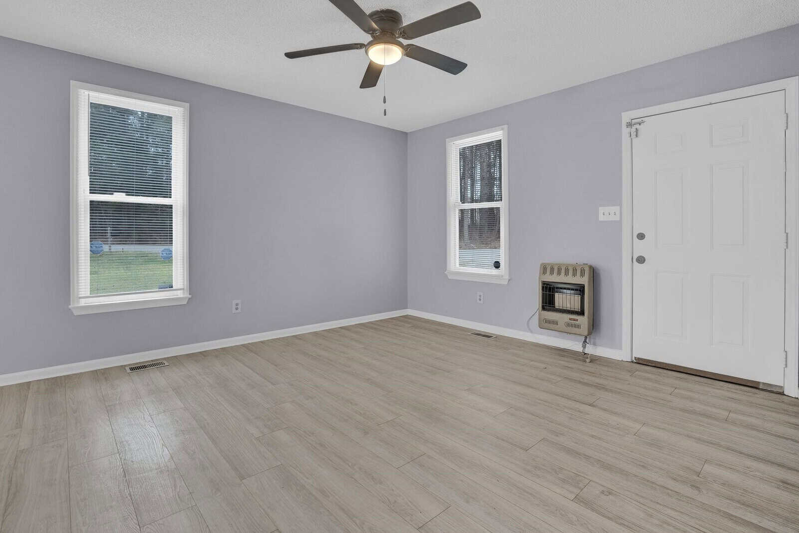 An empty room with light gray walls, laminate flooring, two windows with blinds, a ceiling fan, and a white door.