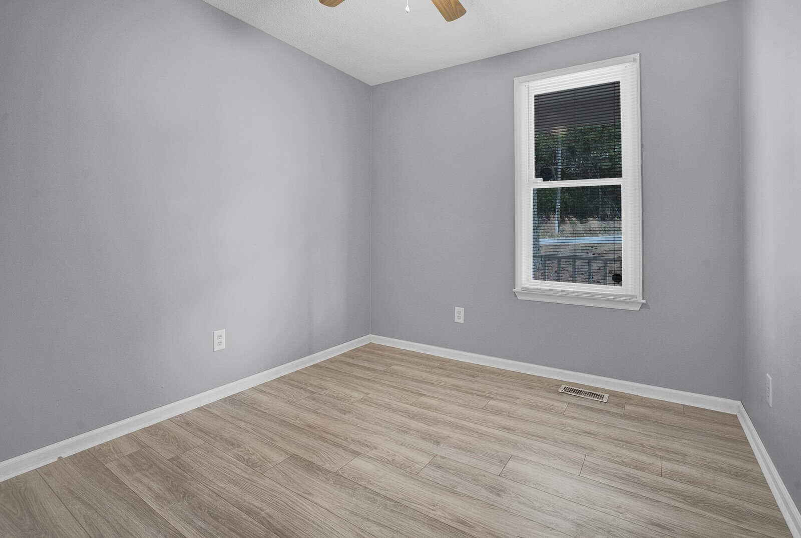 An empty room with gray walls and light wooden laminate flooring, featuring a single window with no treatments.