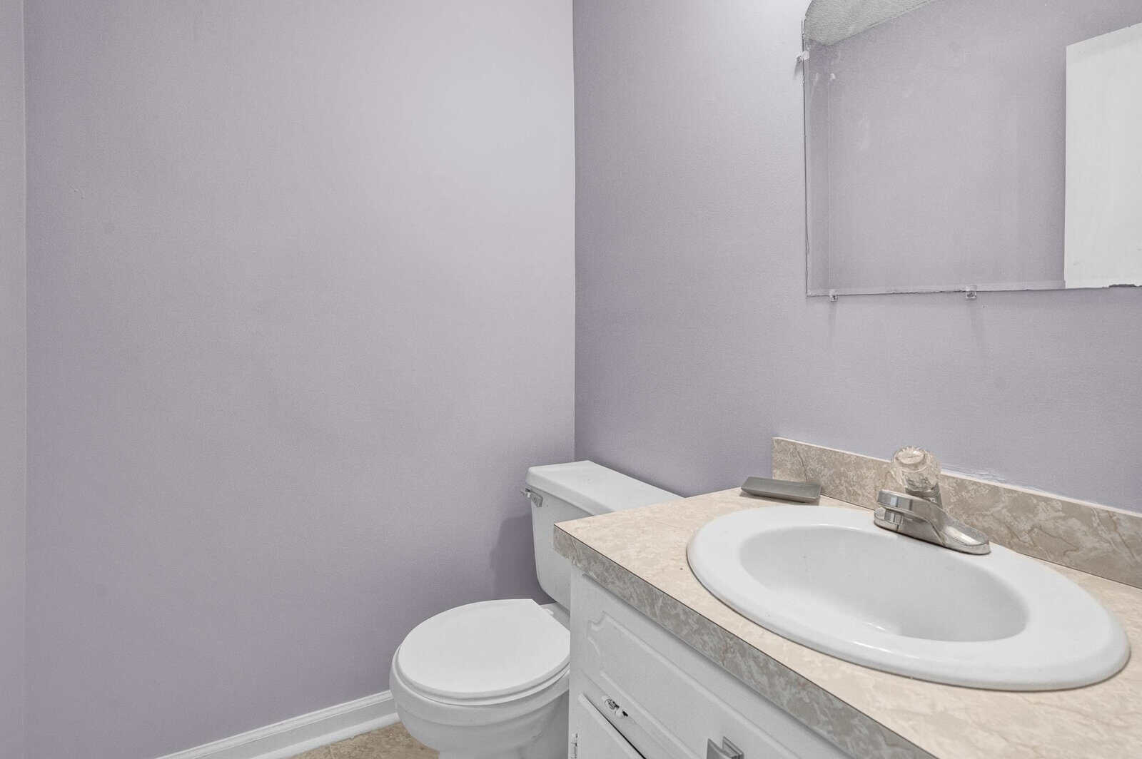 A small bathroom with lavender walls, featuring an incomplete mirror installation above a white sink and vanity, next to a closed toilet.