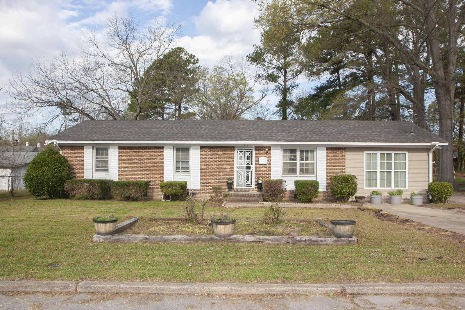 A single-story brick house with white trim and a central white door, flanked by two large windows and situated in front of mature trees under a partly cloudy sky.