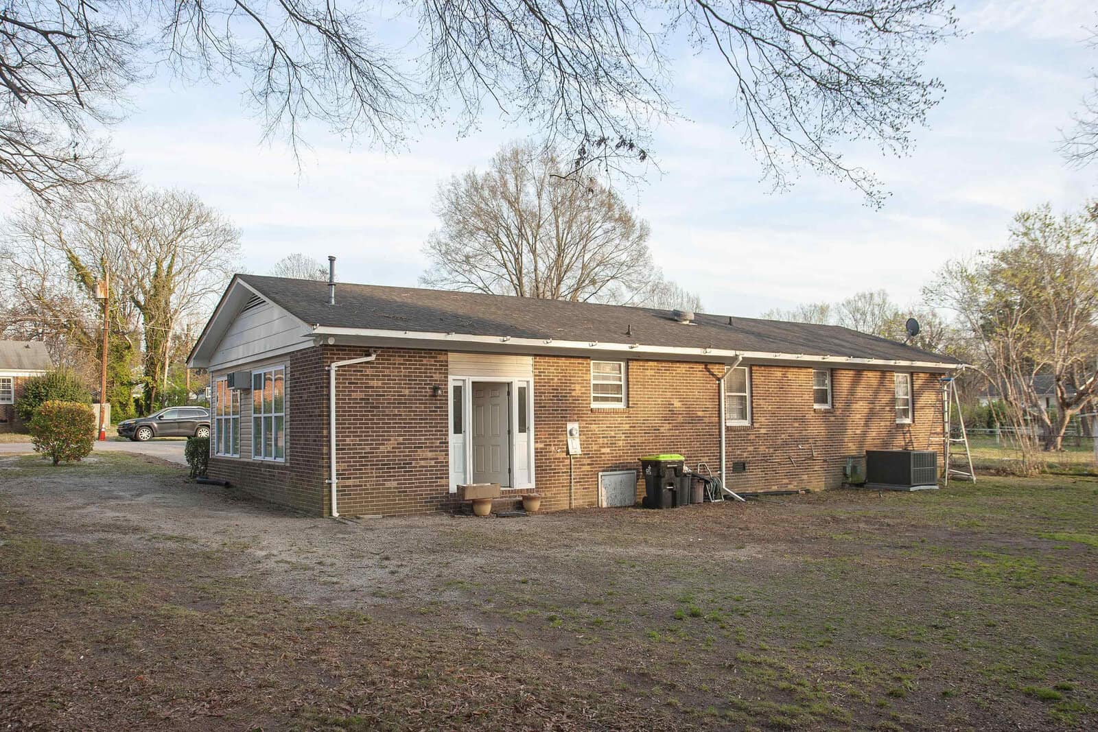 Single-story residential brick building with a white door, visible air conditioning unit, and a screened porch, set against a background of trees with a partial view of a vehicle on the left side. the yard appears to be devoid of grass in several patches.
