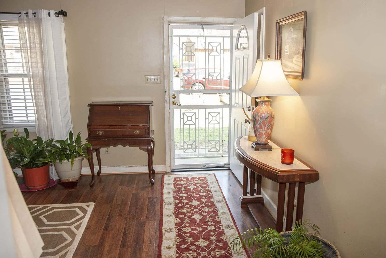 A warmly lit entrance hallway with a wooden floor, featuring an ornate door, a side table with a lamp, a plant, and a small wooden piano against the wall.