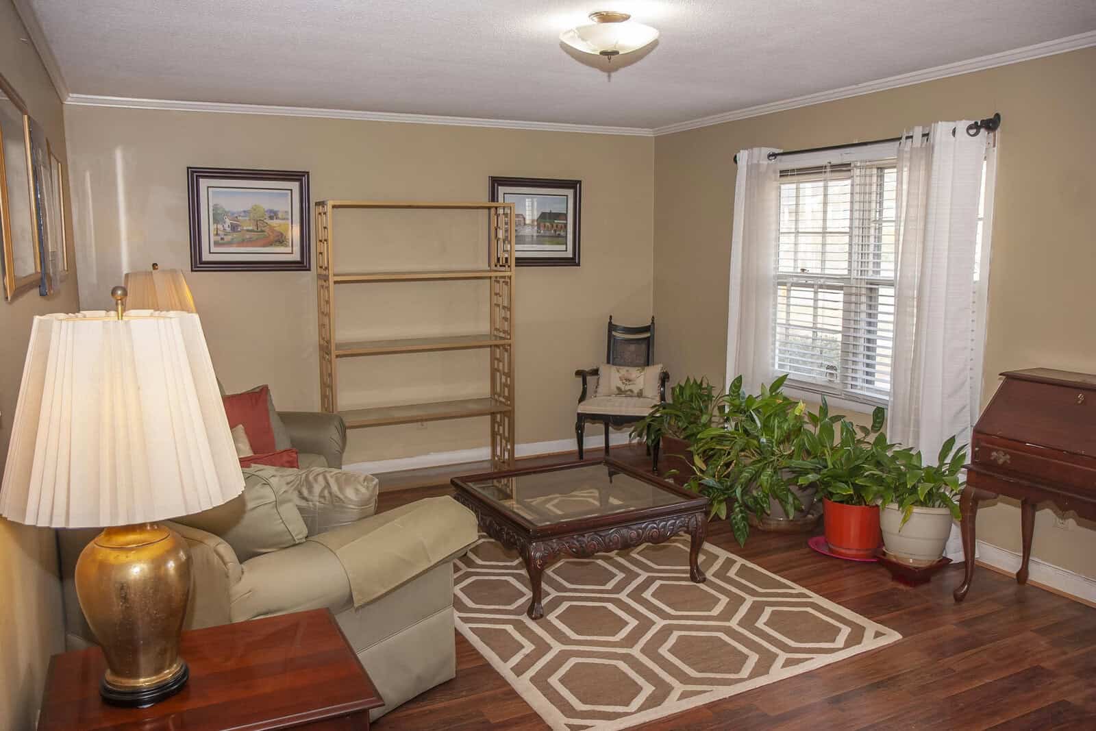 A well-lit, tidy living room with a beige couch, wooden furniture, potted plants, and wall decorations.