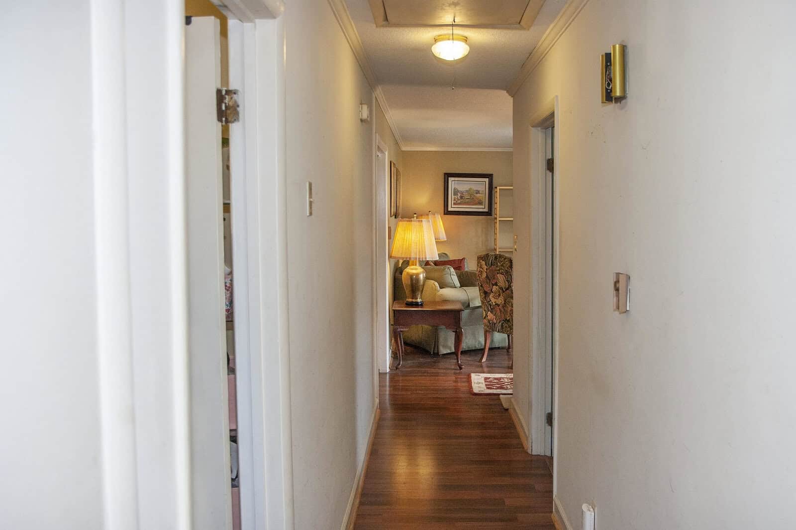 A view down a narrow hallway leading into a cozy room with a lamp, chair, and decorative items, suggesting a warm, lived-in home interior.
