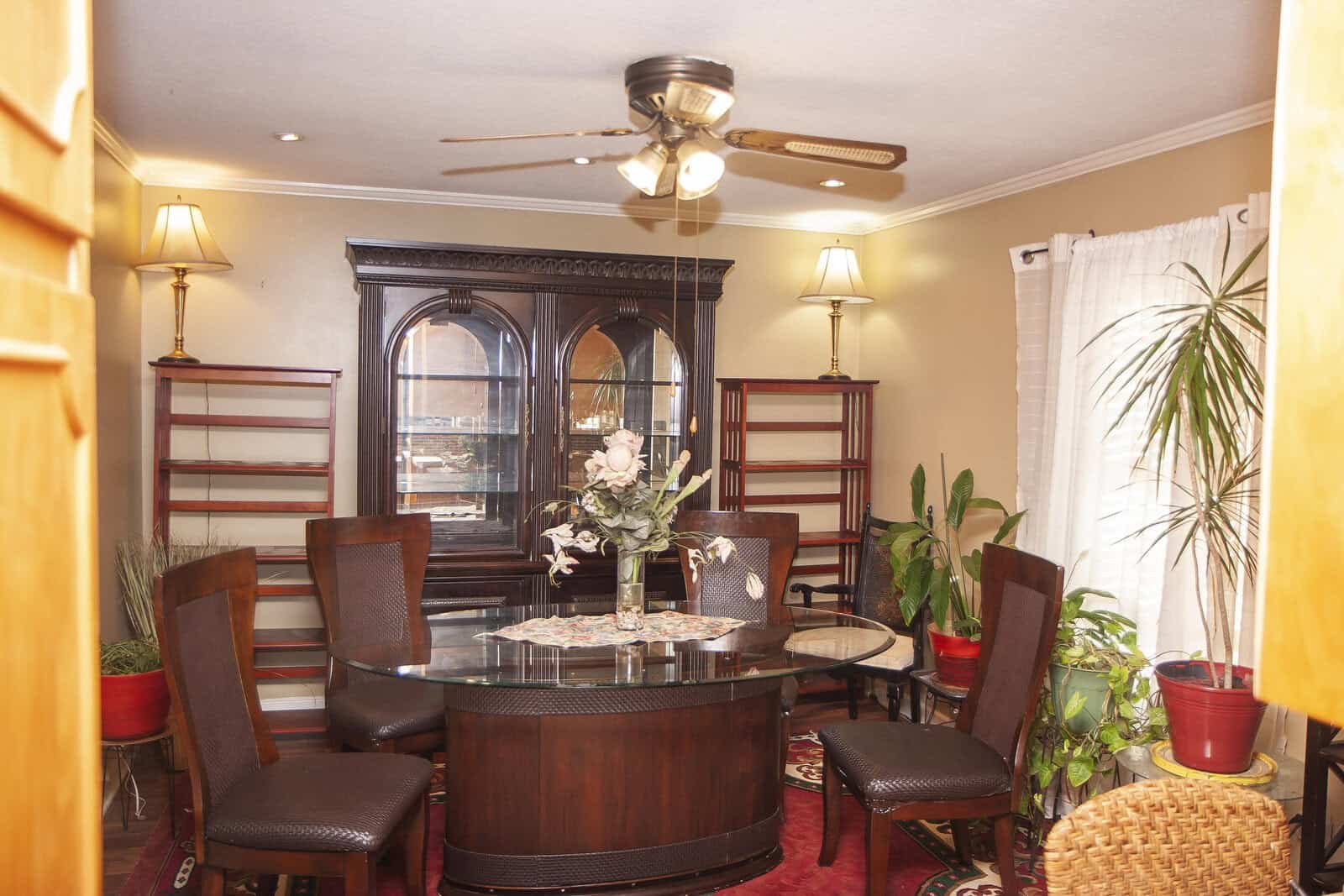 Elegant dining room featuring a round wooden table with matching chairs, a large hutch, and decorative plants, complemented by warm lighting and a ceiling fan.