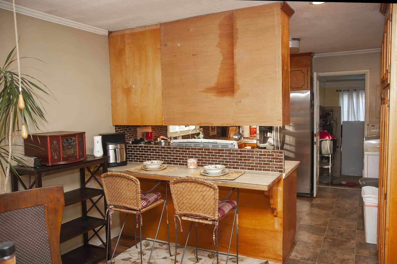A dated kitchen with wooden cabinetry, tiled countertops, and a pair of woven chairs at a breakfast bar.