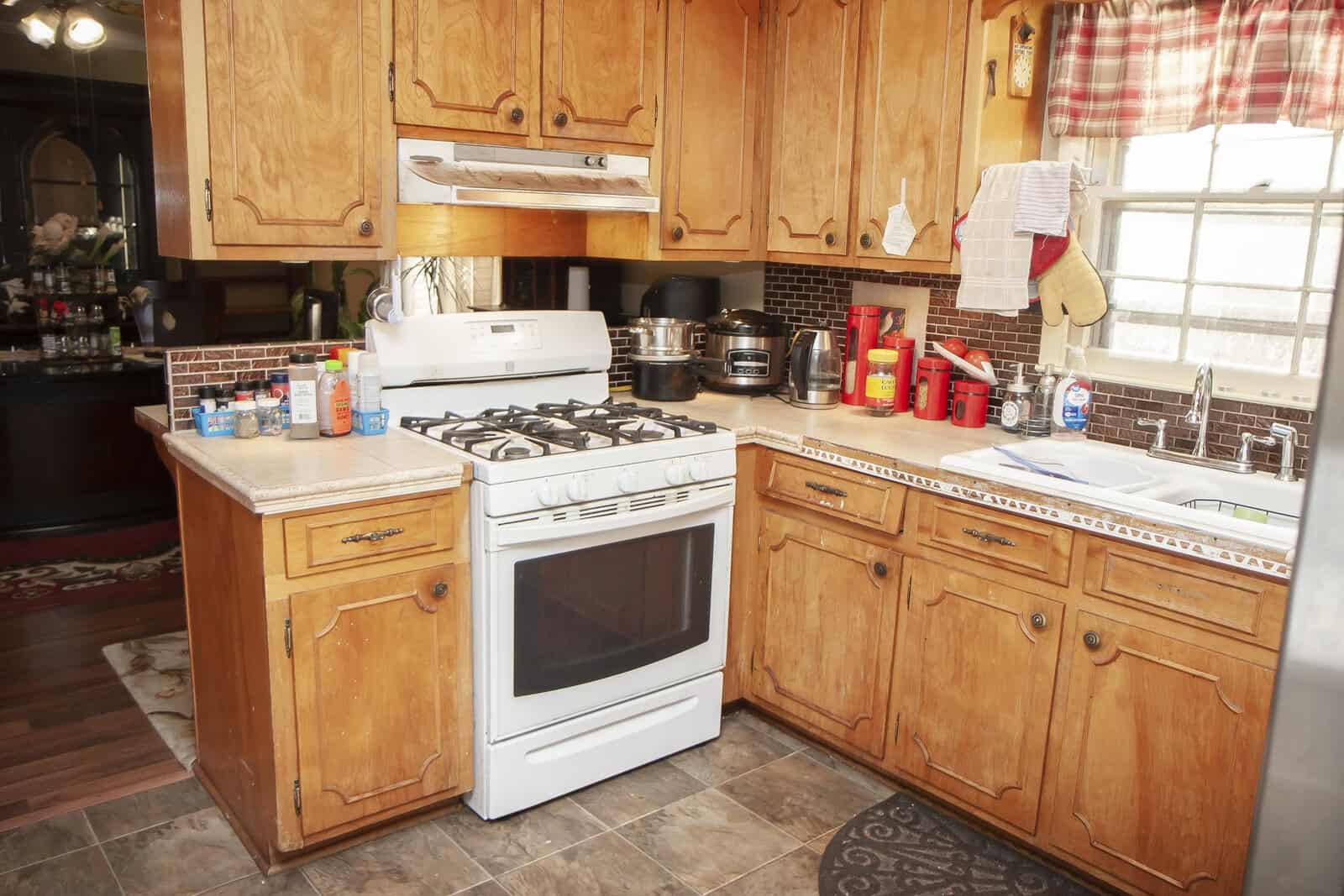 A home kitchen featuring wooden cabinets, a white gas stove, a double sink, and various kitchen utensils and containers scattered on the countertops.