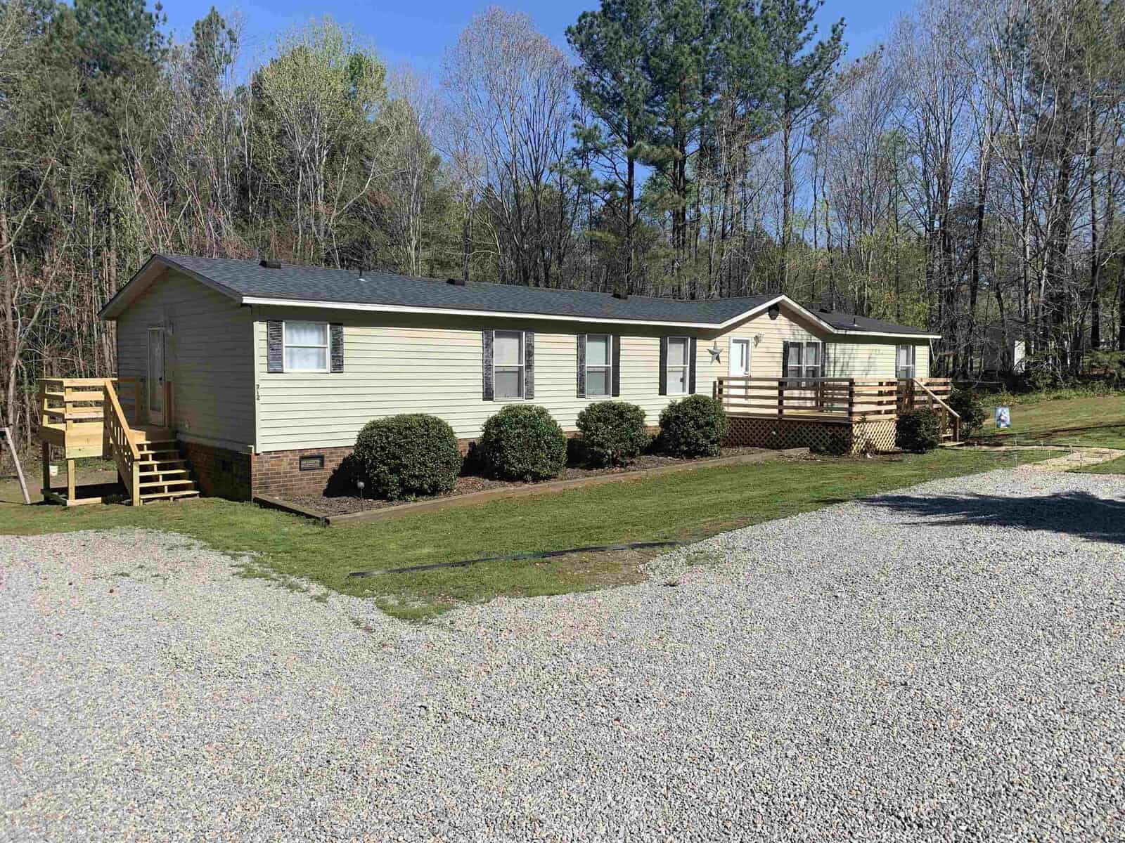 A single-story beige modular home with a brown roof, located at 212 Lydia Lane in Henderson, NC, featuring a wooden front deck and steps, surrounded by a grassy lawn with trees in the