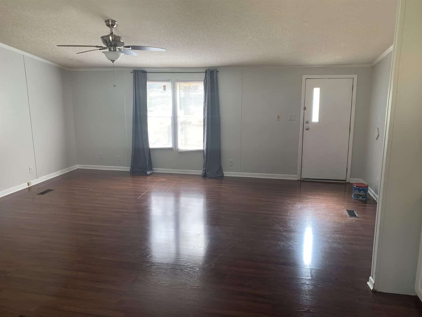 An empty room with glossy hardwood flooring, white walls, a ceiling fan, and a single window draped with dark curtains.