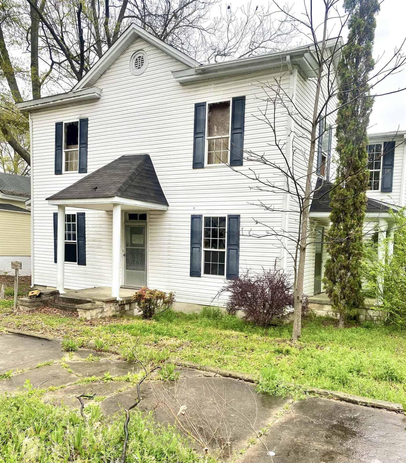 A two-story white house with black shutters, featuring an arched entryway, surrounded by greenery and situated next to a driveway at 146 Burwell Avenue, Henderson NC.