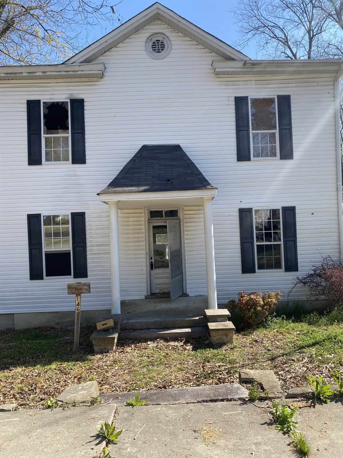 A two-story white house with a black gabled portico, missing window panes, and signs of disrepair.