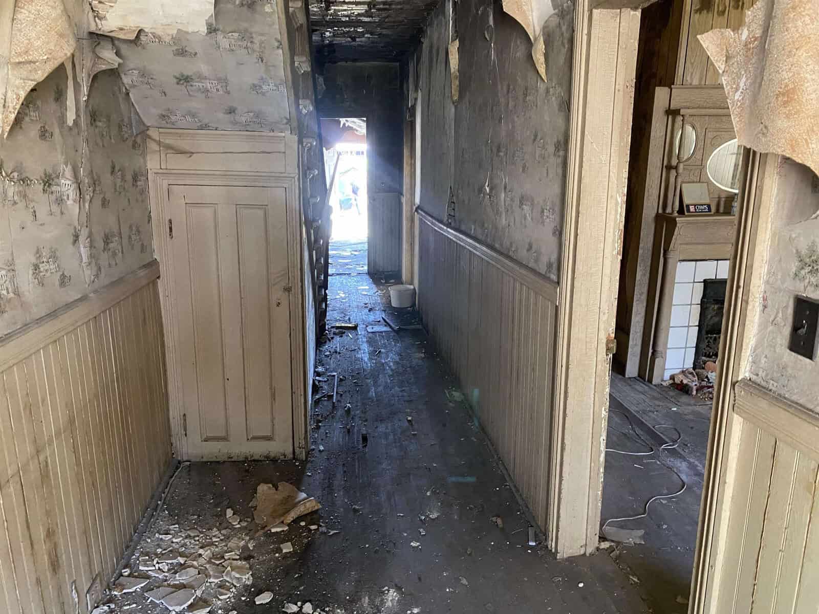 Interior corridor of a dilapidated building showing signs of decay and neglect, with peeling wallpaper, scattered debris, and a partially illuminated end from an exterior light source.