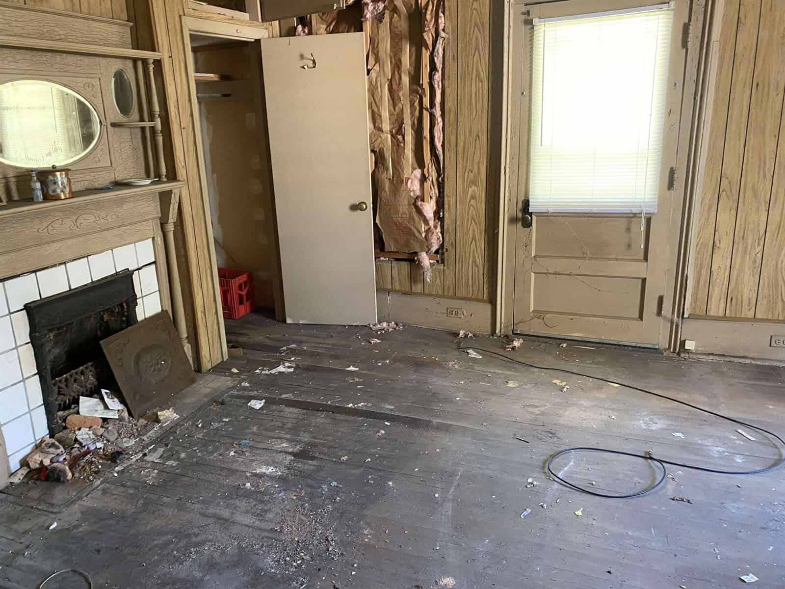 An old, neglected room with a fireplace, wood paneling, and debris scattered across the hardwood floor.