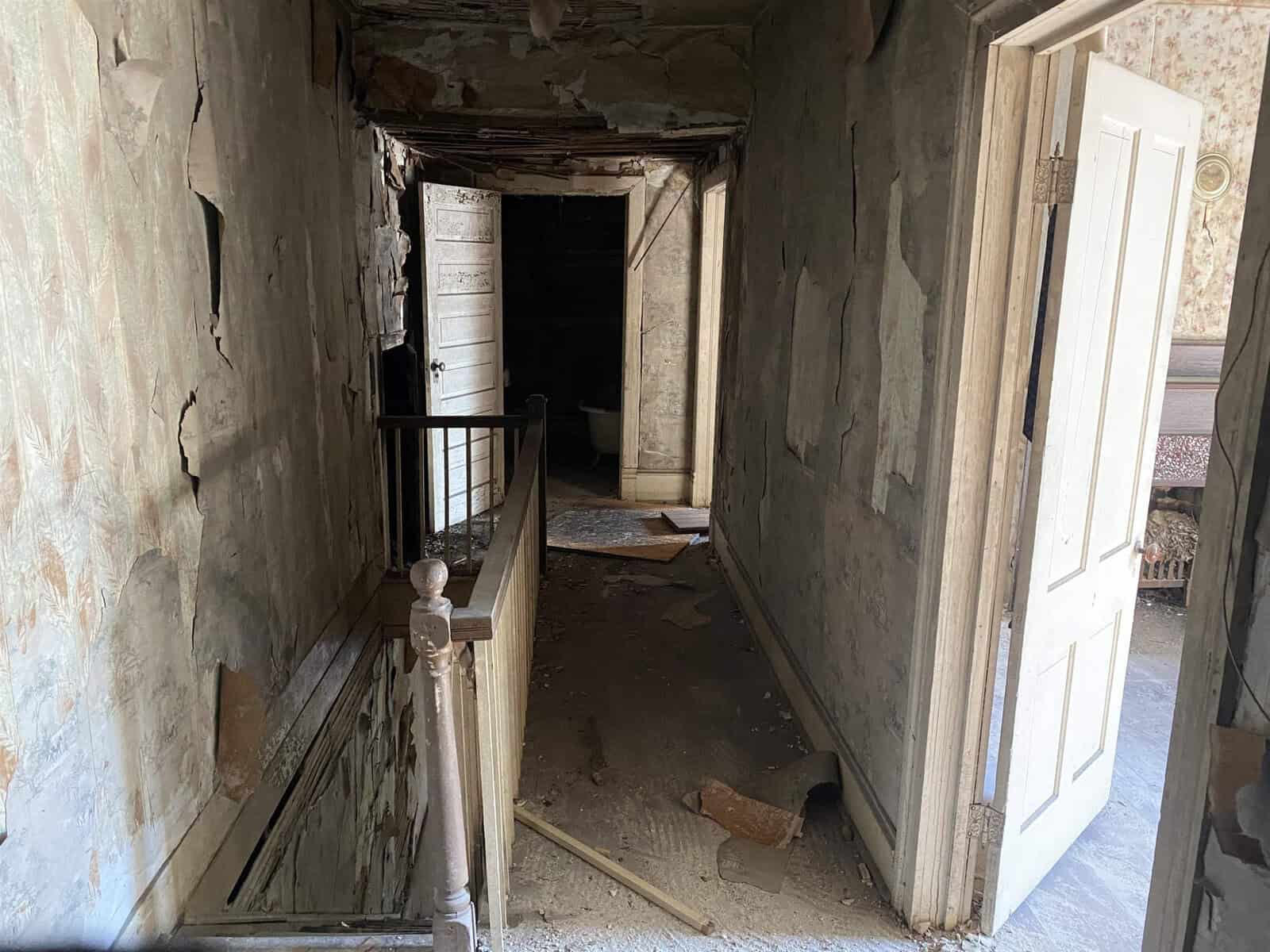 A dilapidated interior hallway of an old building, featuring peeling wallpaper, damaged walls, and debris on the floor, with a staircase railing on the left.