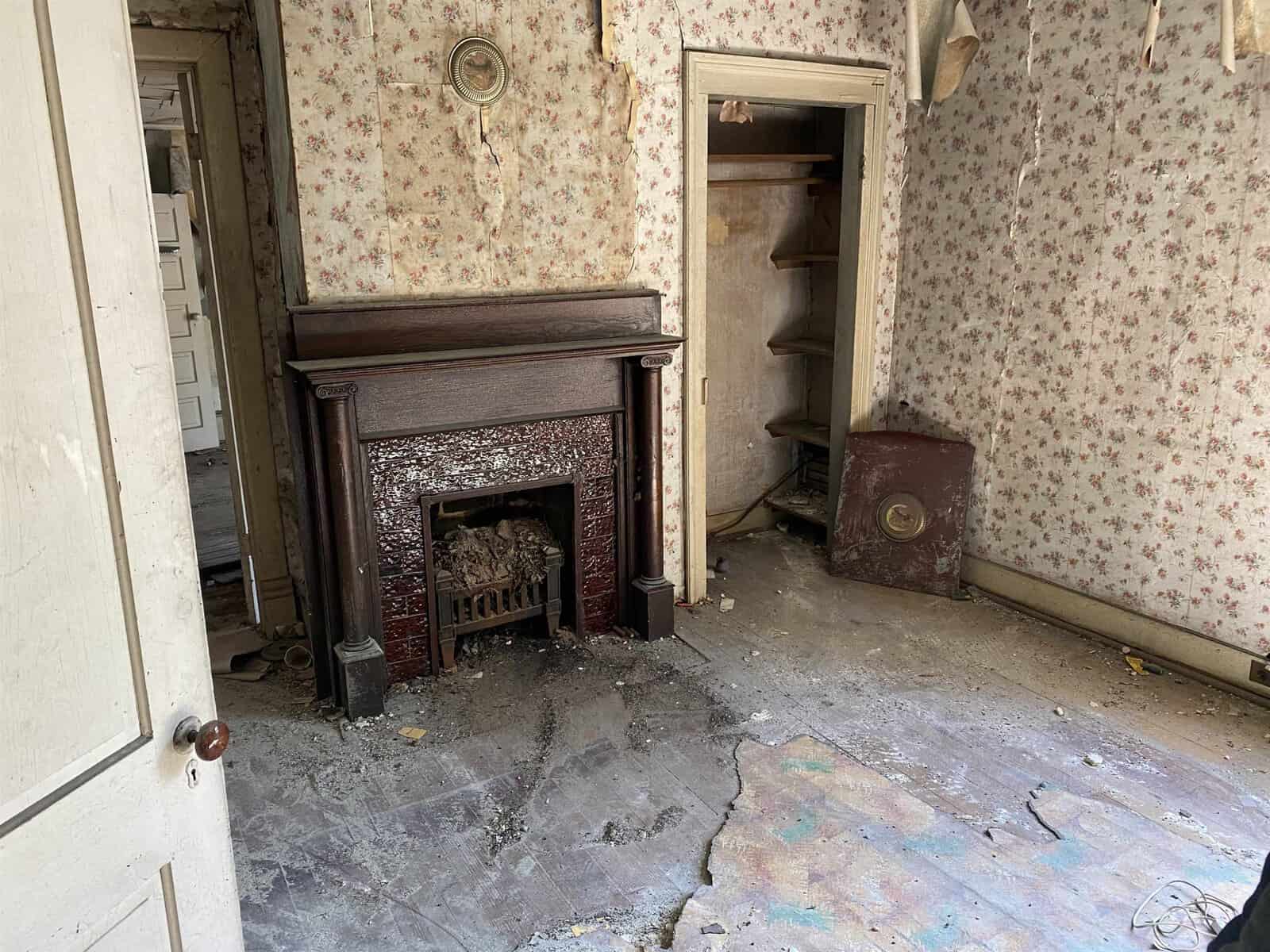 An old, abandoned room with a floral wallpaper, featuring a dusty fireplace and a damaged floor with debris. a corner built-in shelving unit and an antique heating stove are visible to the right.
