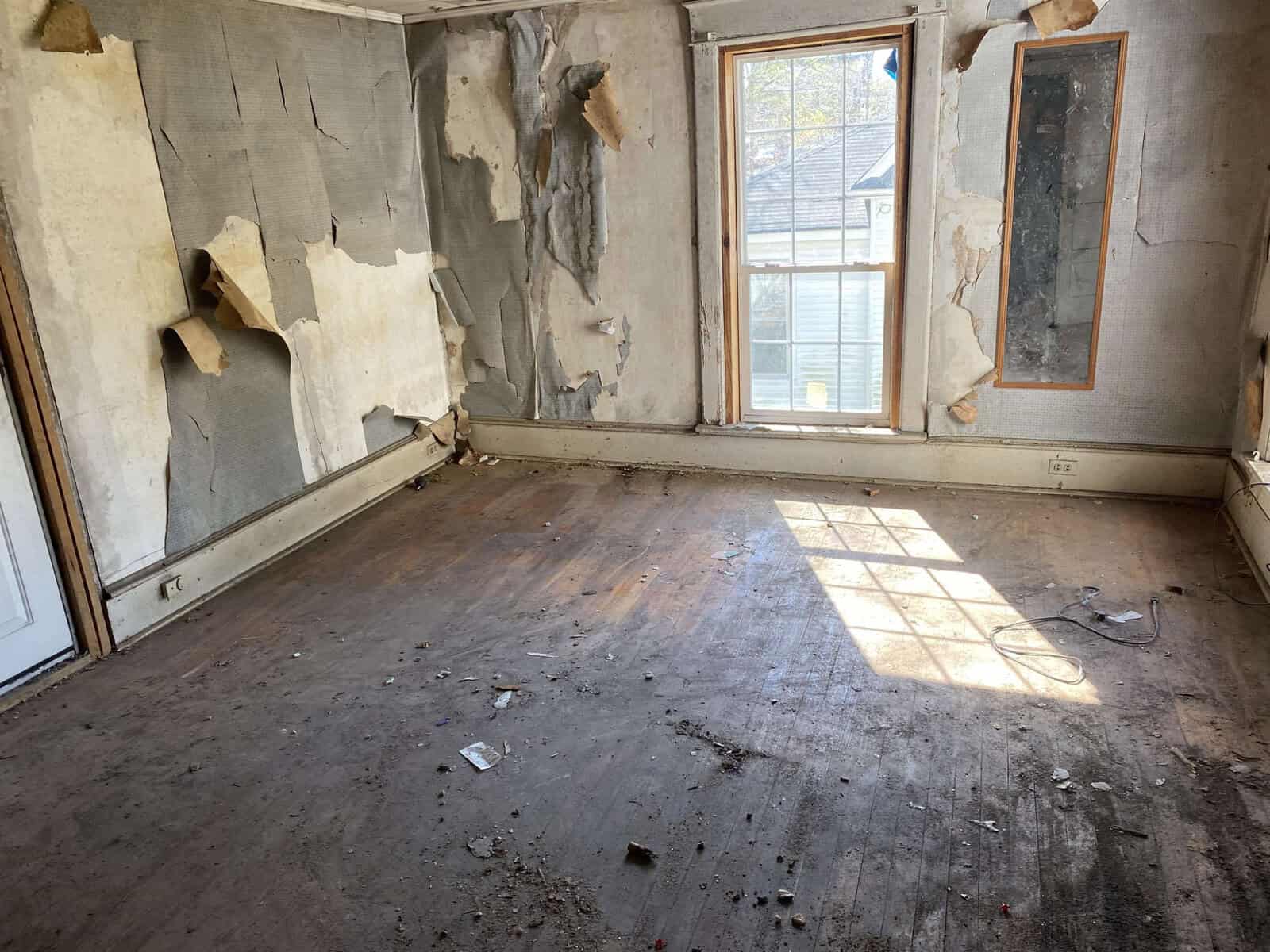A room with peeling wallpaper, a bare wooden floor, and sunlight coming through a window with torn curtains.