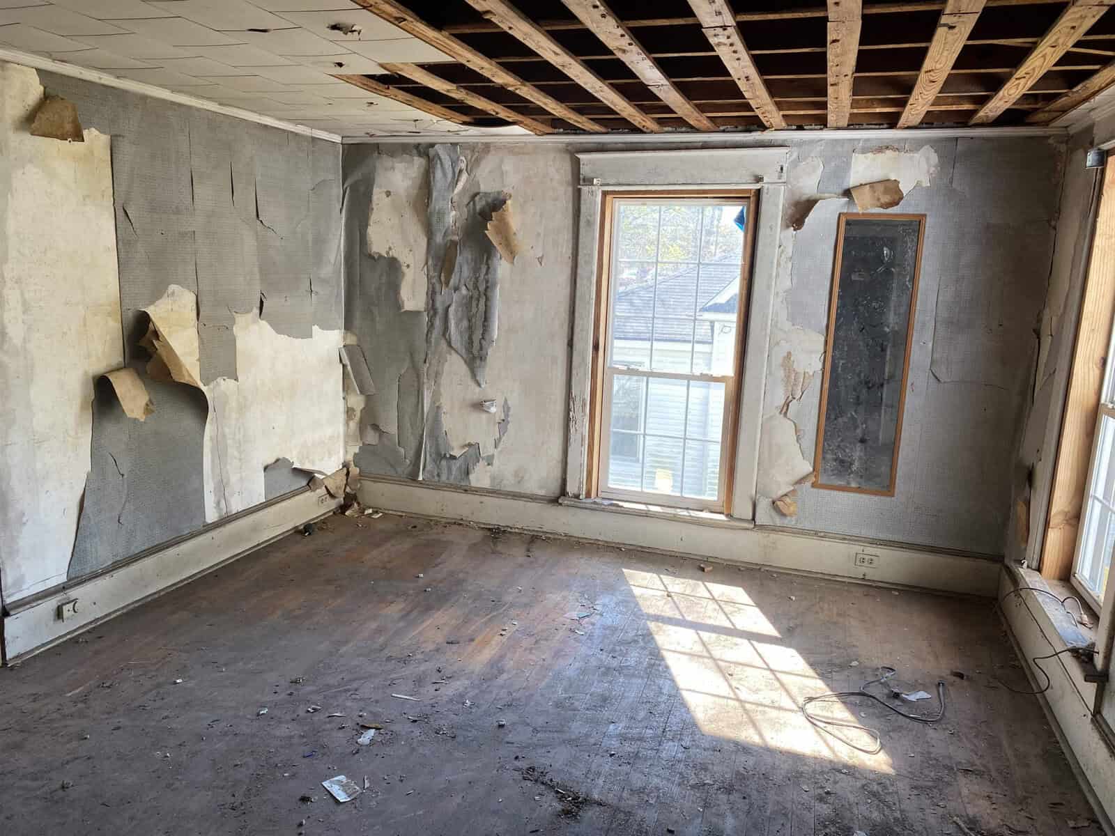 A dilapidated room with torn wallpaper, exposed ceiling joists, and a worn wooden floor, with sunlight streaming through a window.