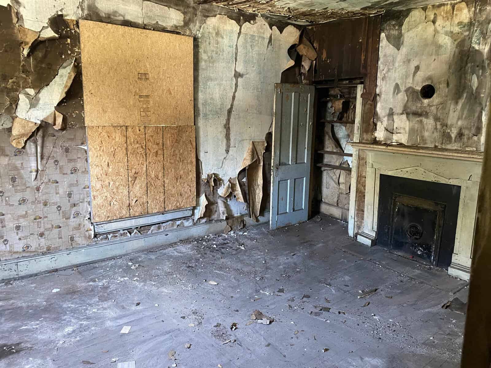 An abandoned room with peeling walls, a boarded-up window, a damaged door leaning against the wall, and a fireplace.