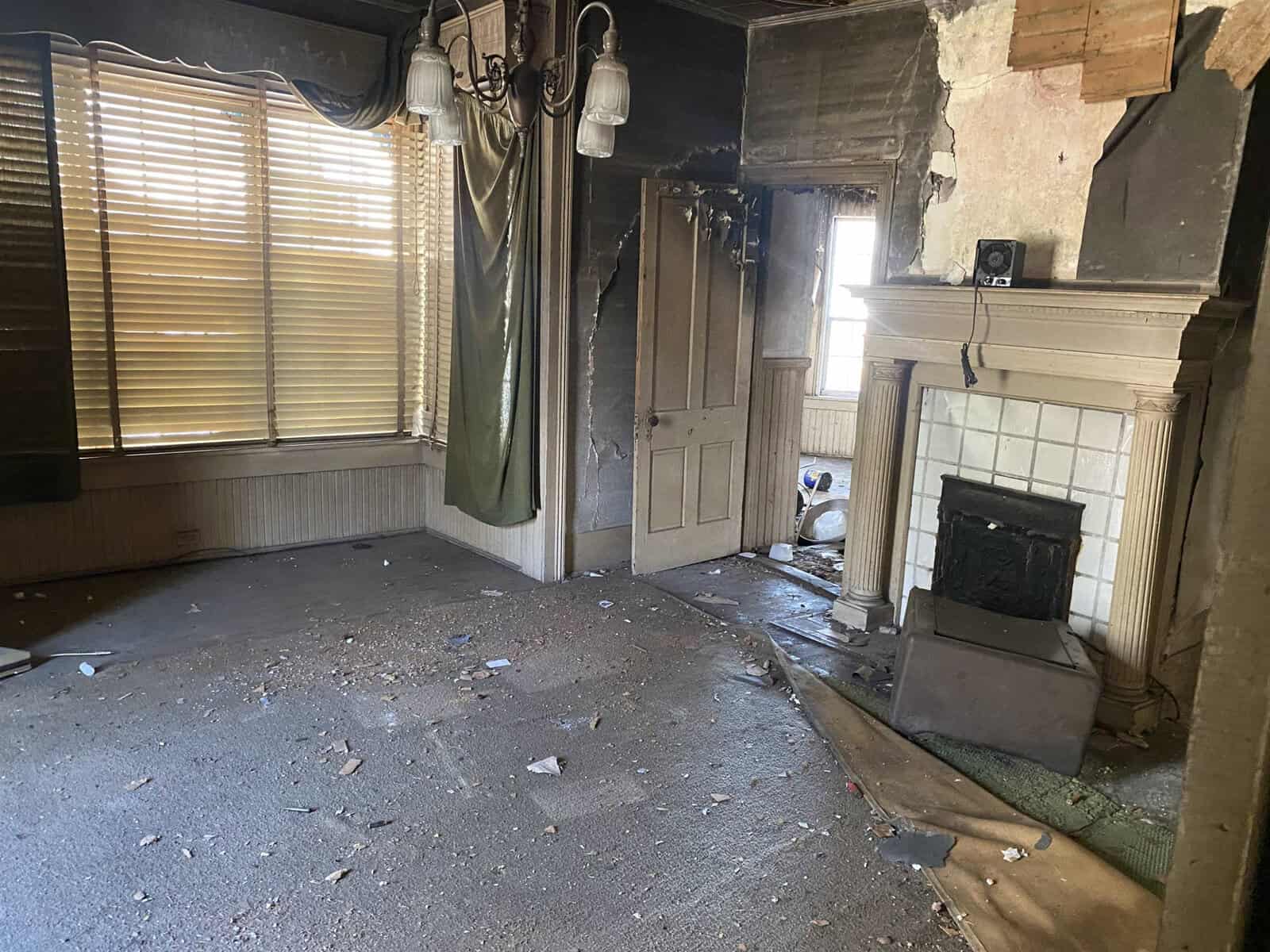 Interior of an abandoned room with damaged walls, a deteriorated fireplace, and debris scattered on the floor, with closed blinds covering the windows.