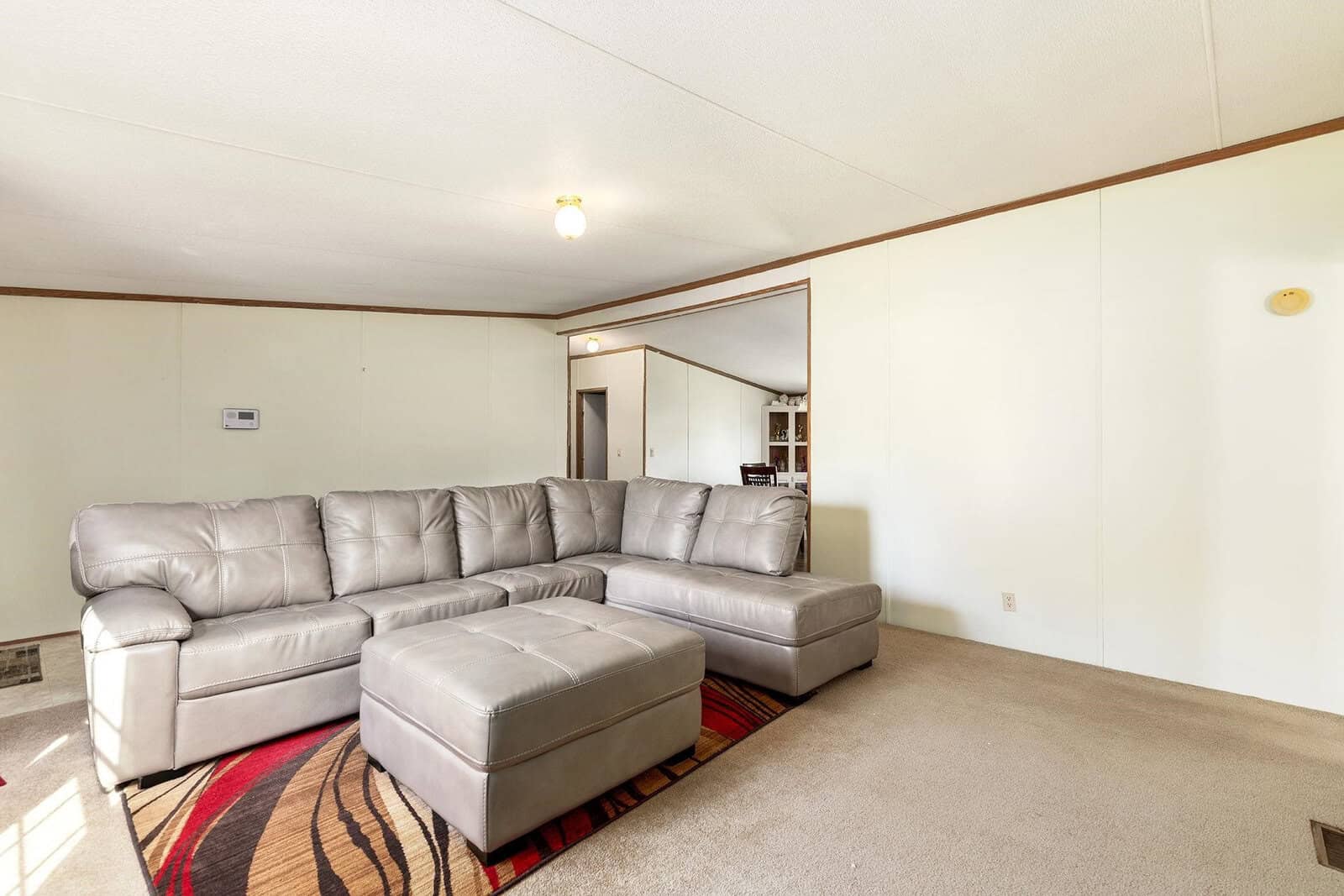 A large, grey sectional sofa with an ottoman on a patterned rug in an empty living room with beige walls and carpet flooring.