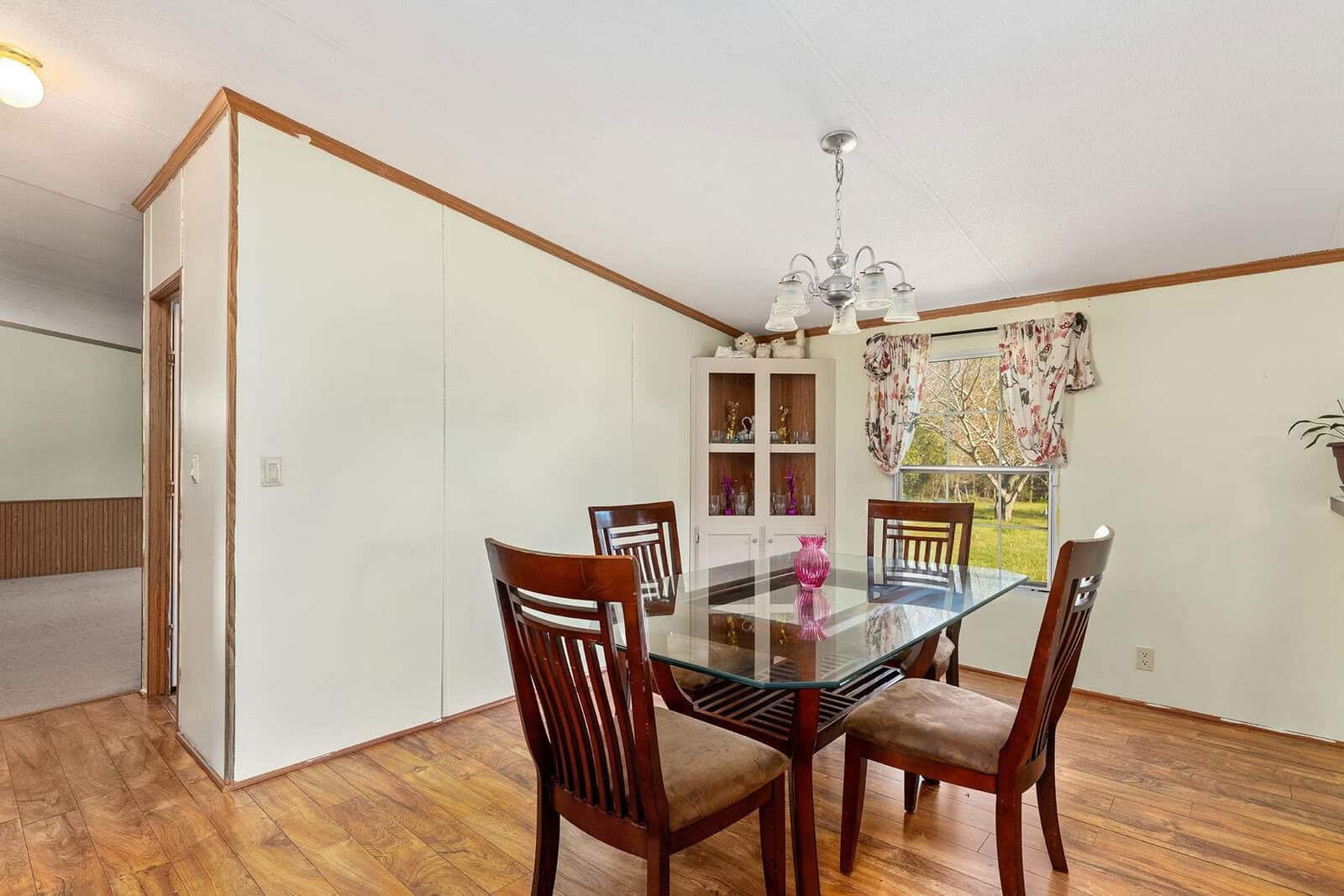 A modest dining room with a wooden table set, four chairs, a white shelf, and a chandelier, featuring hardwood floors and a large window with floral curtains.