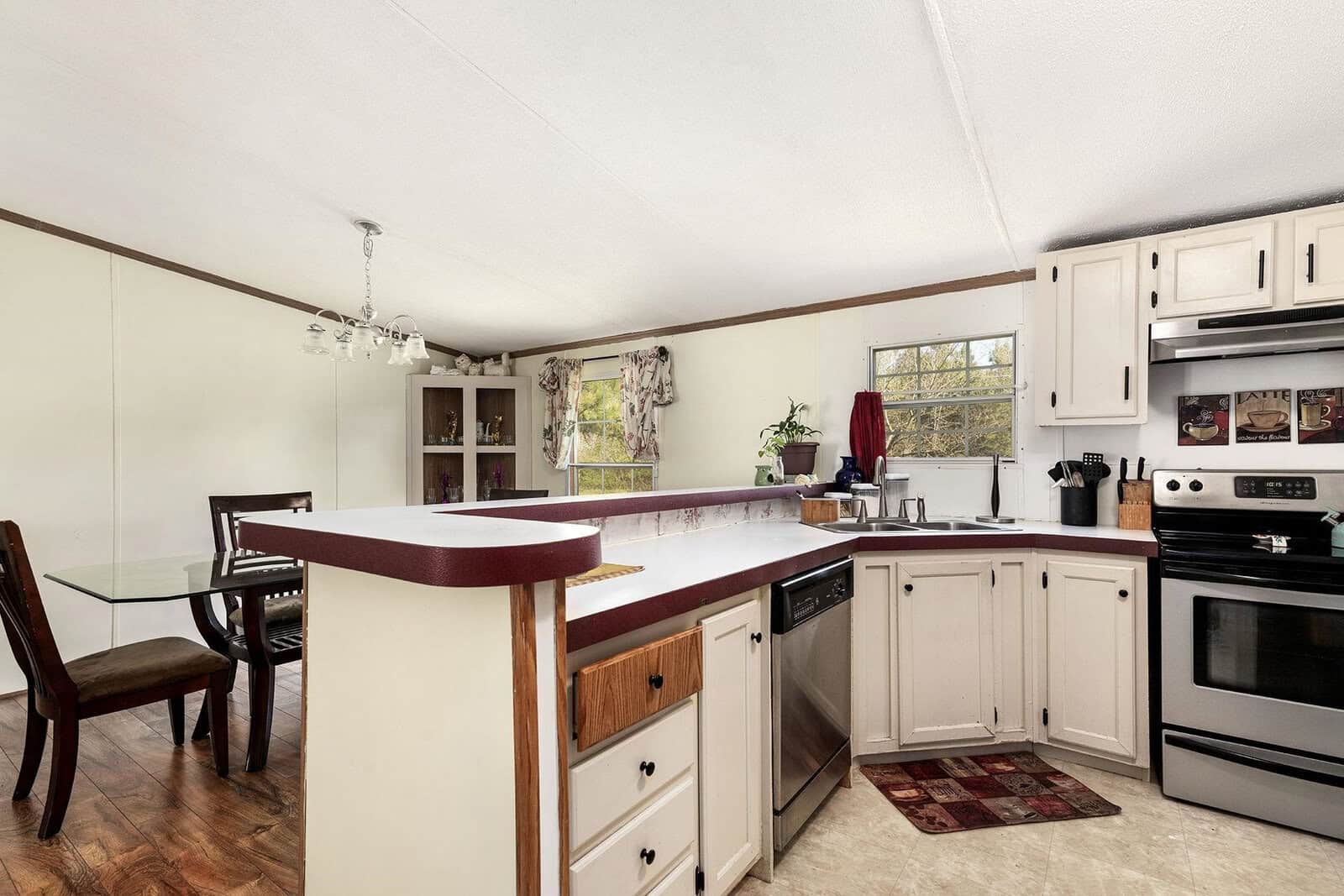 A modest, well-lit kitchen with white cabinetry and laminate countertops, featuring a range of appliances and a small dining area with a glass-top table and chairs.