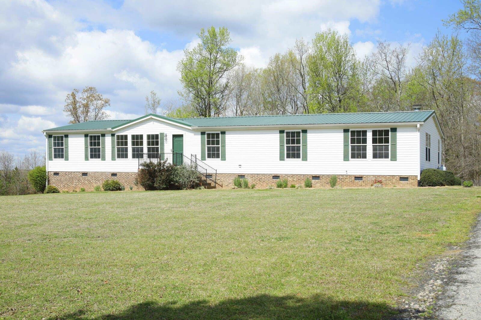 A single-story white modular home with a green roof and partial brick foundation, positioned on a grassy plot with a backdrop of trees under a partly cloudy sky.