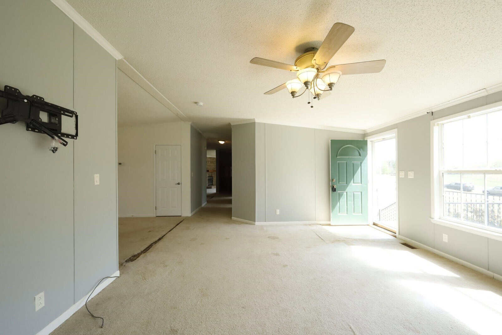 Empty residential room with wall-to-wall carpeting, a ceiling fan with lights, and a sliding glass door leading to a balcony. there is a mounted tv bracket on the left wall.