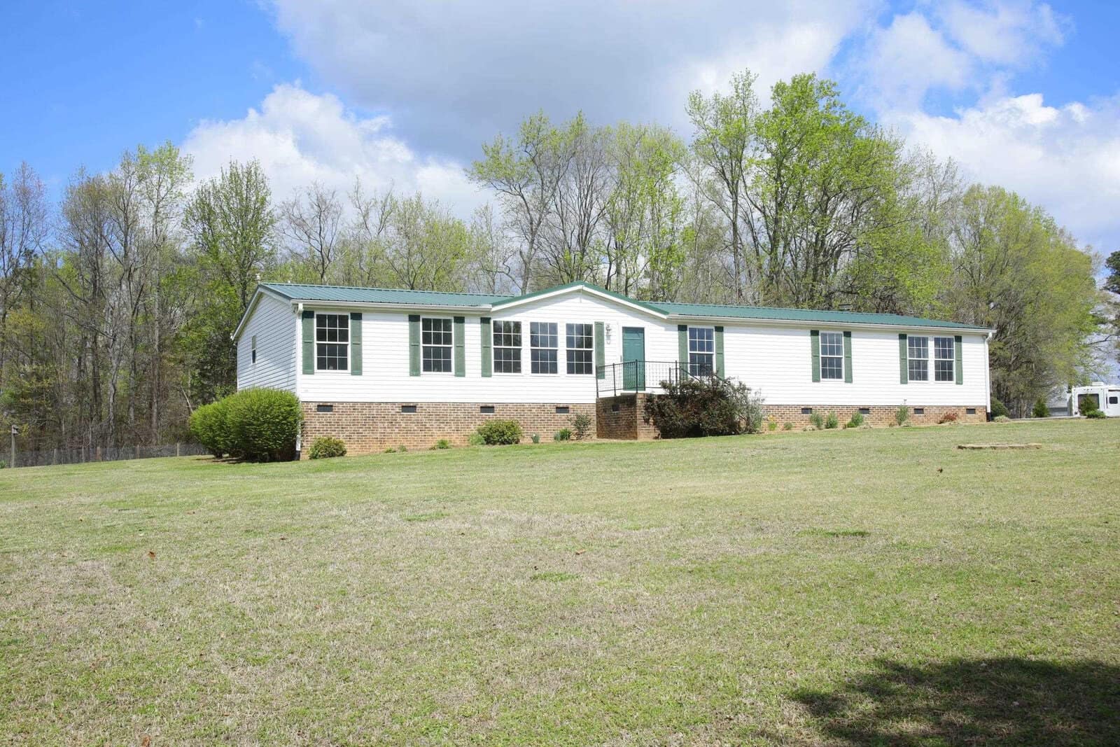 A single-story white modular home with a green roof, set against a backdrop of a clear blue sky and surrounded by a well-kept lawn with a few scattered trees.