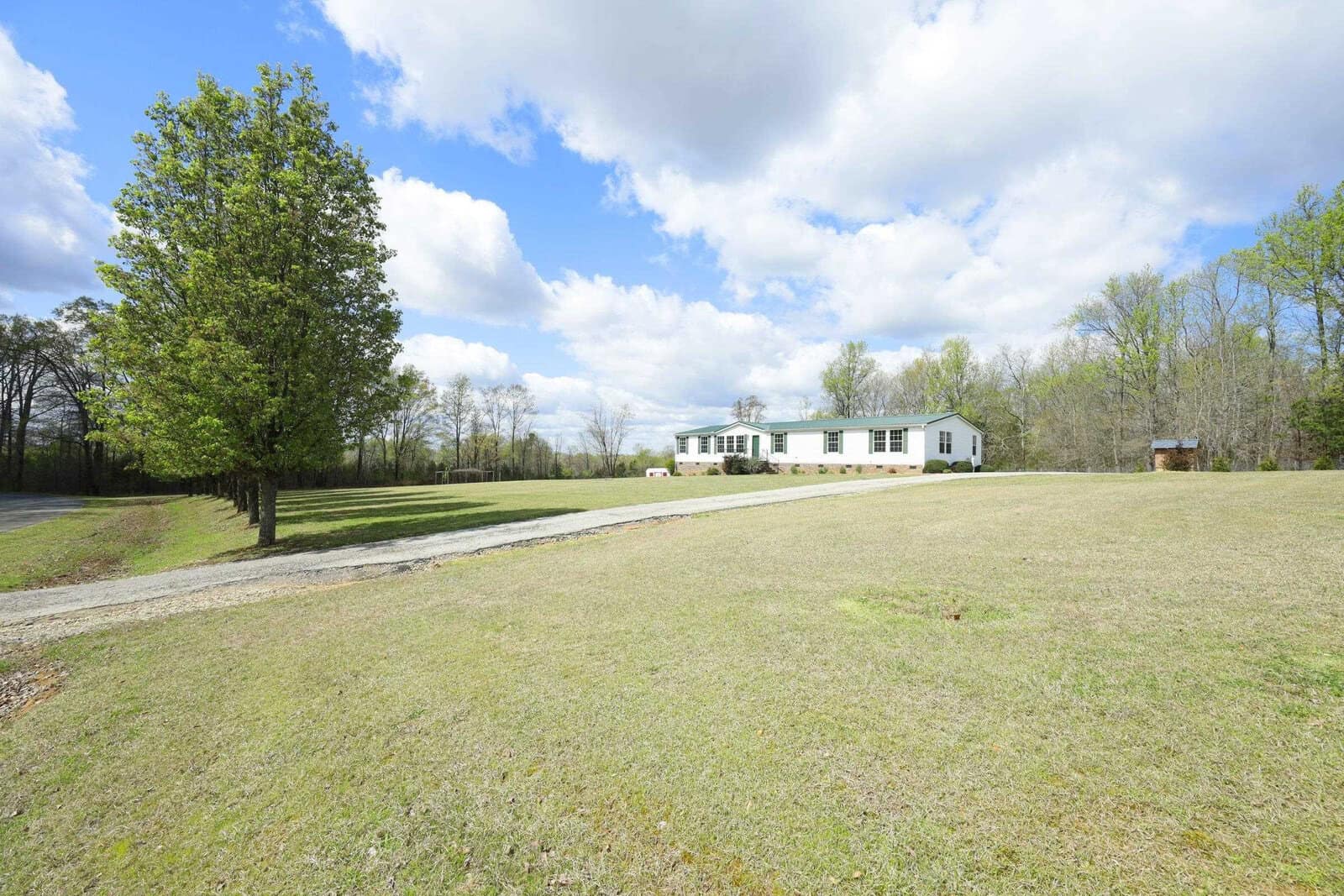 A single-story house with a white facade and green roof situated in a large open yard with scattered trees under a partly cloudy sky.