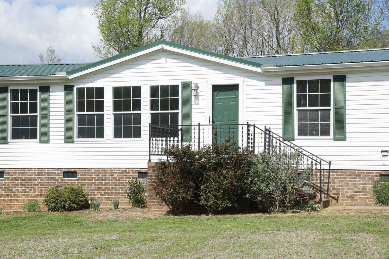 A white manufactured home with green shutters, a brick foundation, and a small porch with metal railings.