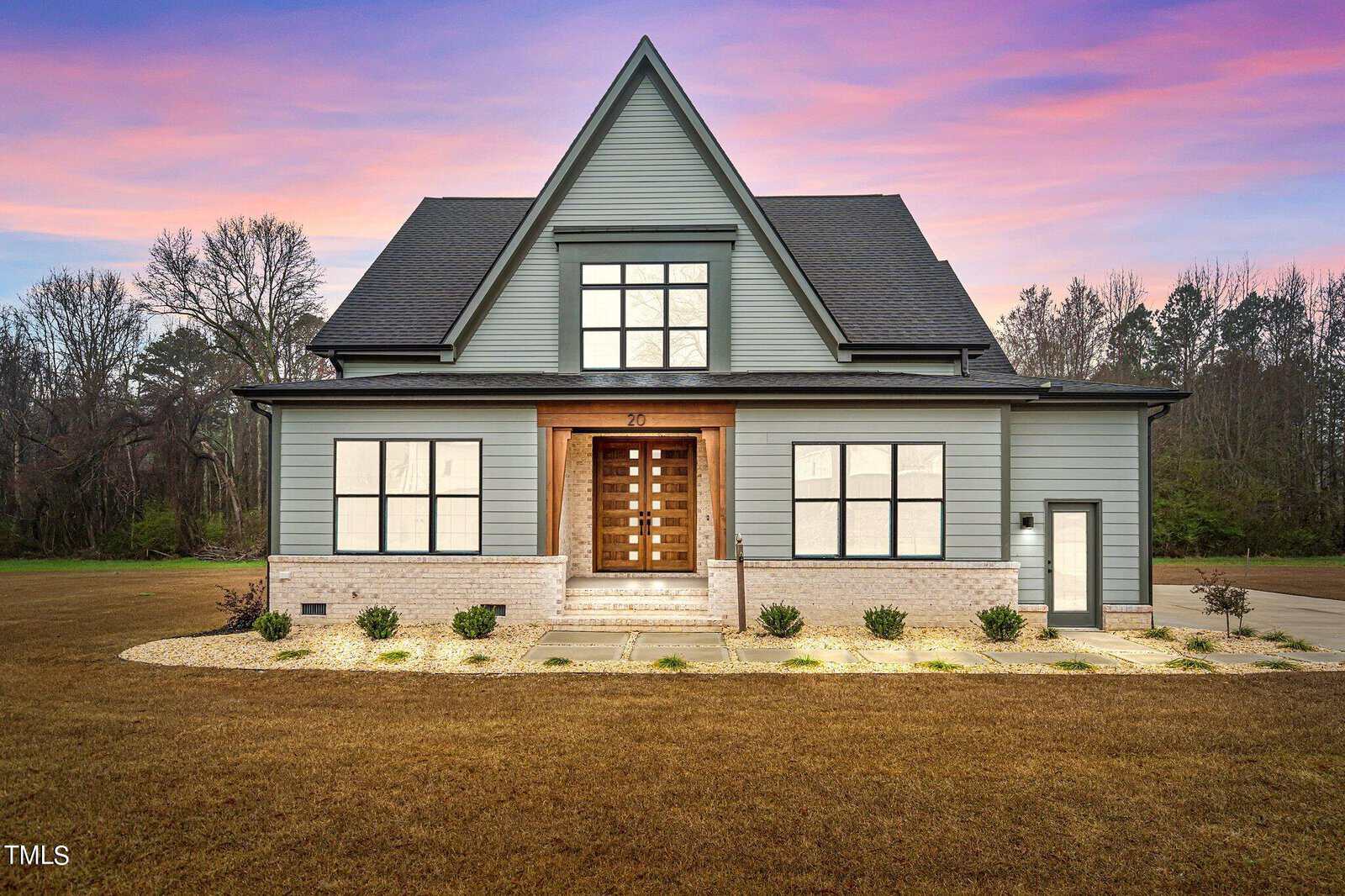 Modern two-story house with gray siding, large windows, and a prominent peaked roof, set on a spacious lot with a manicured lawn, landscaping, and a dramatic sunset sky in the background.