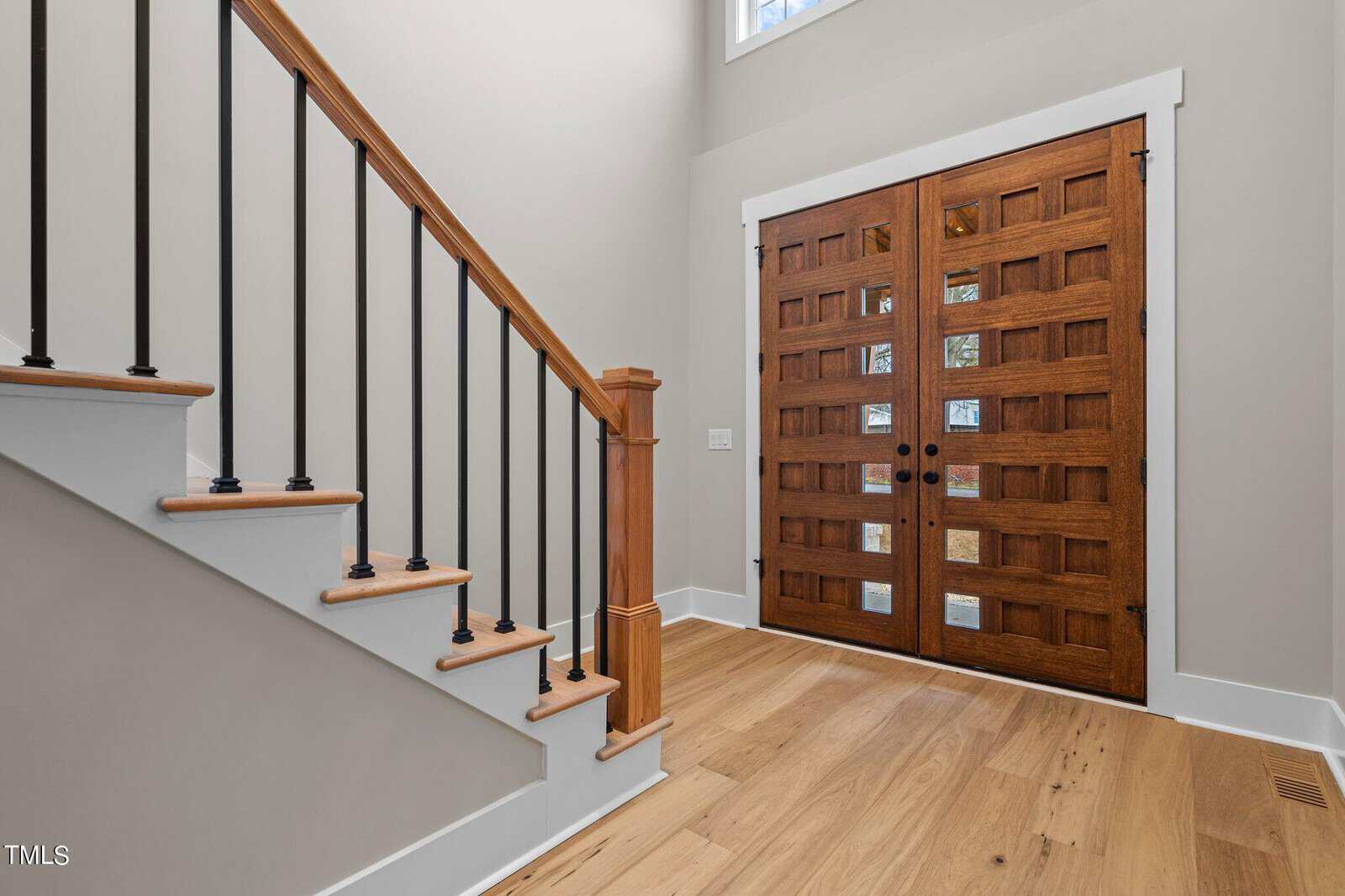 A bright entryway featuring wooden double doors with glass panels, light wood flooring, and a staircase with black balusters and wooden handrail. Neutral-colored walls and natural light create an inviting atmosphere.