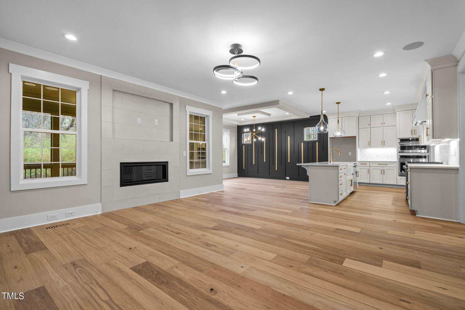 Open-concept living area with light wood floors, modern fireplace, and large windows. Adjacent kitchen features white cabinets, gold accents, pendant lights, and a dark accent wall with built-in appliances.