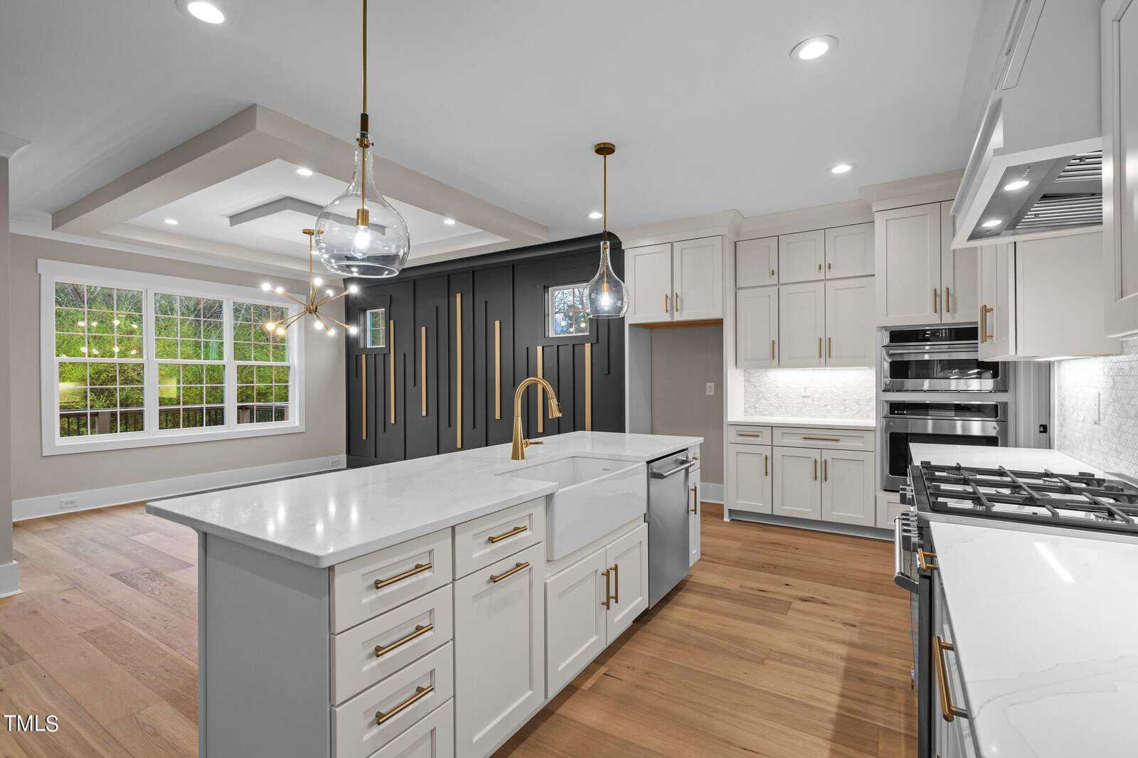 Modern kitchen with white cabinets, gold hardware, a large island with a sink, stainless steel appliances, light wood floors, and a dark accent wall. Open layout connects to a bright dining area with large windows.