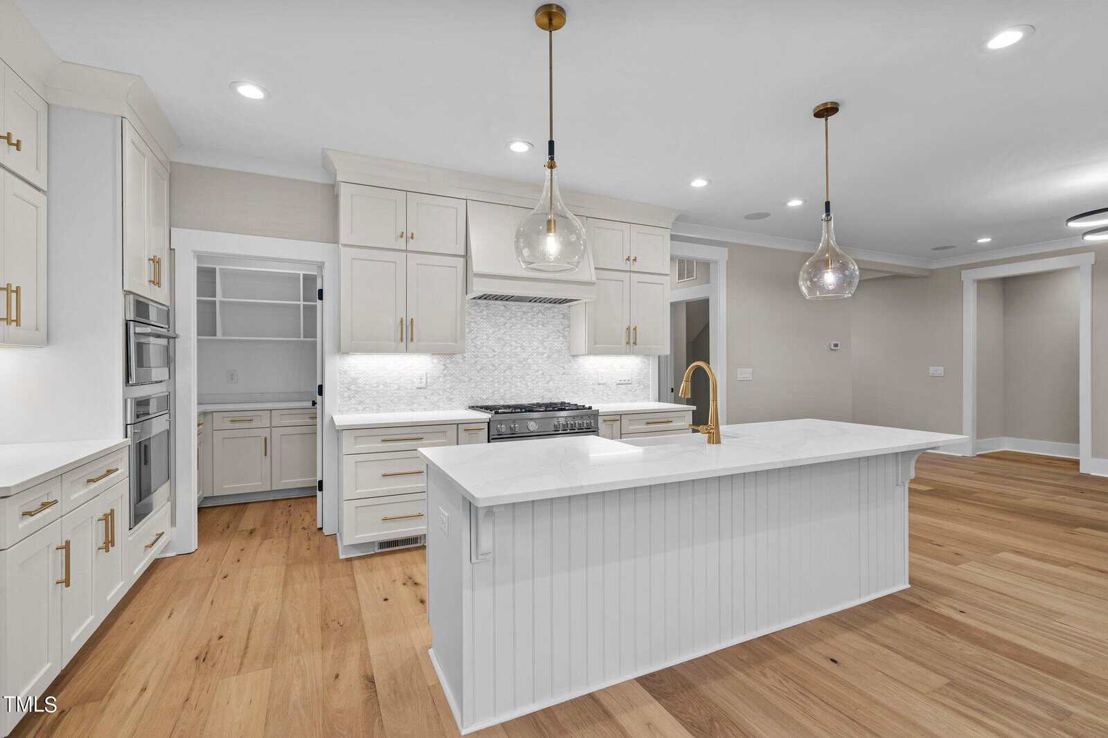Modern kitchen with white cabinets, gold hardware, a white island with a farmhouse sink, pendant lights, light wood floors, gas range with hood, built-in oven, and a walk-in pantry in the background.