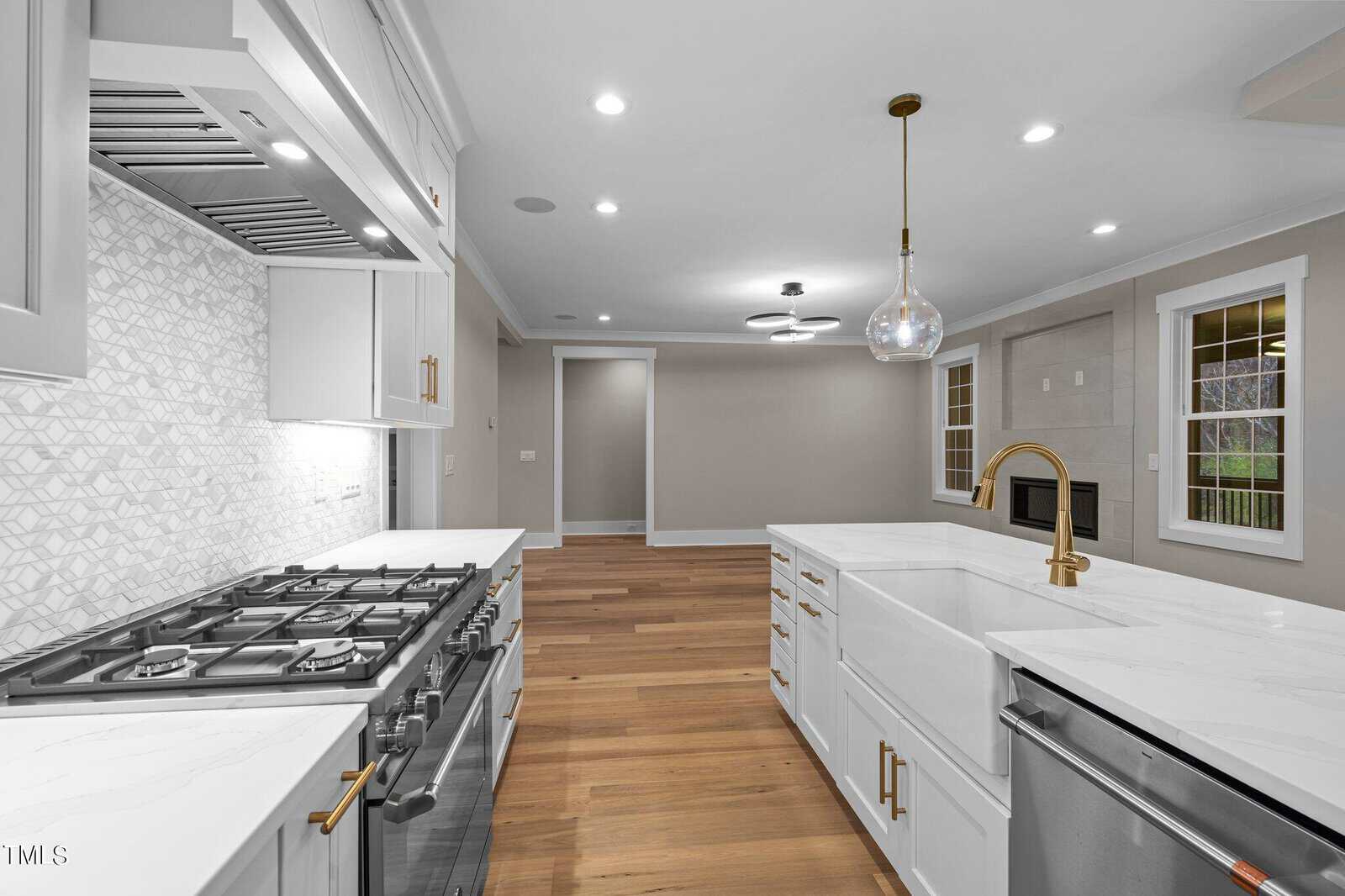 Modern kitchen with white cabinets, marble countertops, stainless steel gas stove and dishwasher, gold hardware, farmhouse sink, pendant light, and wood flooring. Neutral walls and a window are visible in the background.