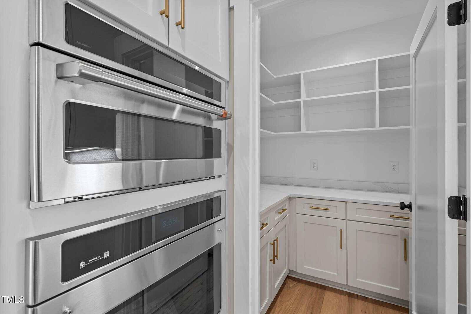 Modern kitchen with built-in stainless steel double oven on the left, adjacent to a white pantry with gold hardware, marble countertops, open shelves, and wood flooring.
