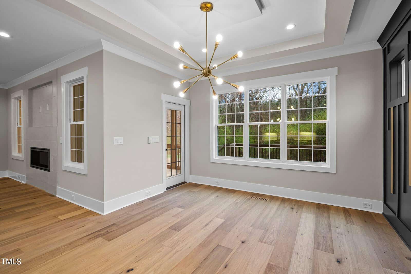 Modern, empty room with light wood floors, large windows, beige walls, a glass door, tray ceiling, and a gold sputnik chandelier. Outdoor greenery is visible through the windows.