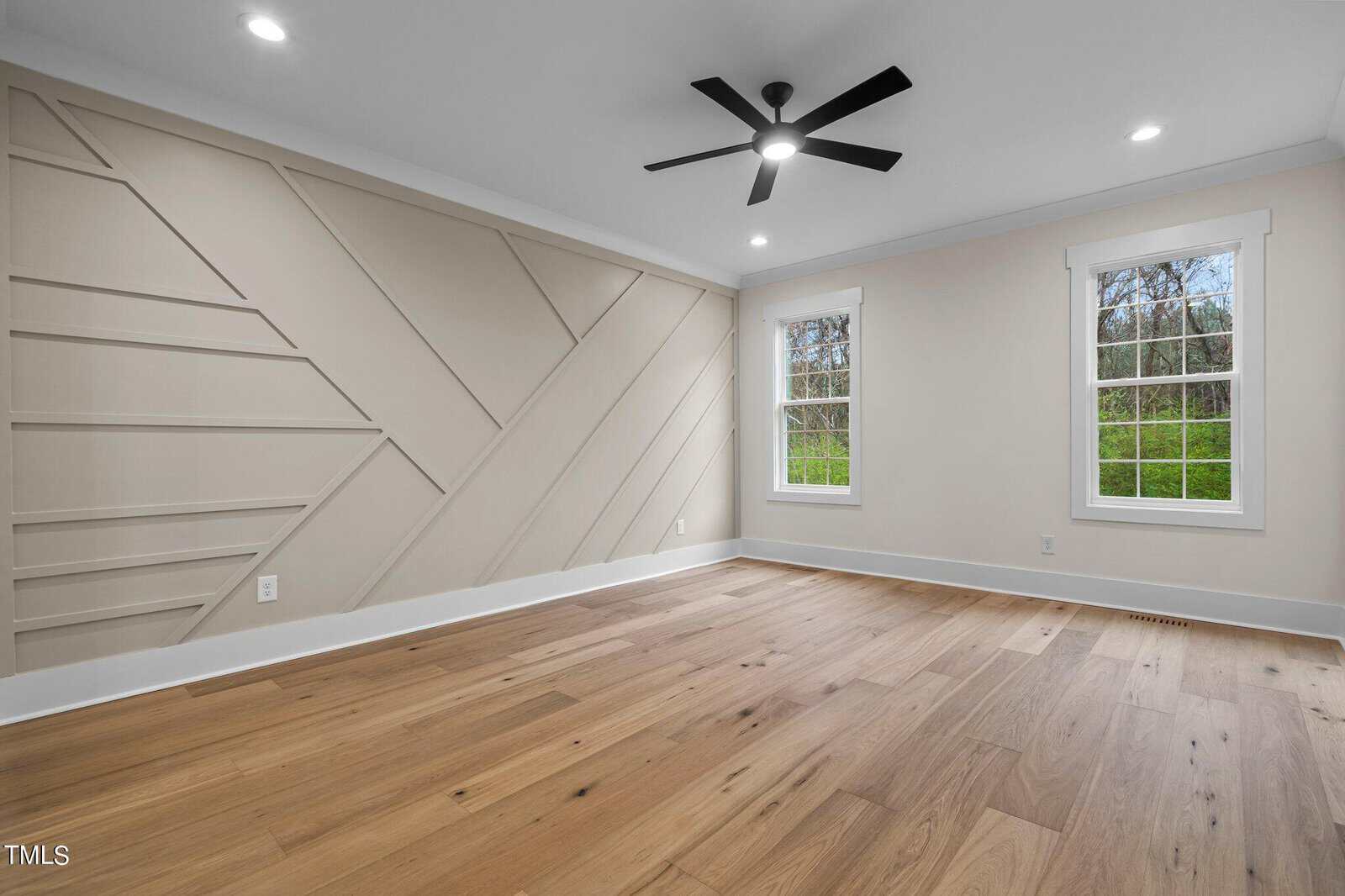 A modern, empty room with light wood floors, two large windows, white walls, geometric accent wall paneling, recessed lighting, and a black ceiling fan. Natural light streams in through the windows.
