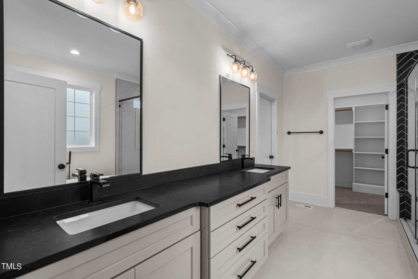 Modern bathroom with dual sinks, black countertops, two mirrors, and light fixtures. A walk-in closet with shelves is visible through an open door on the right. The walls are light-colored and the floor is tiled.