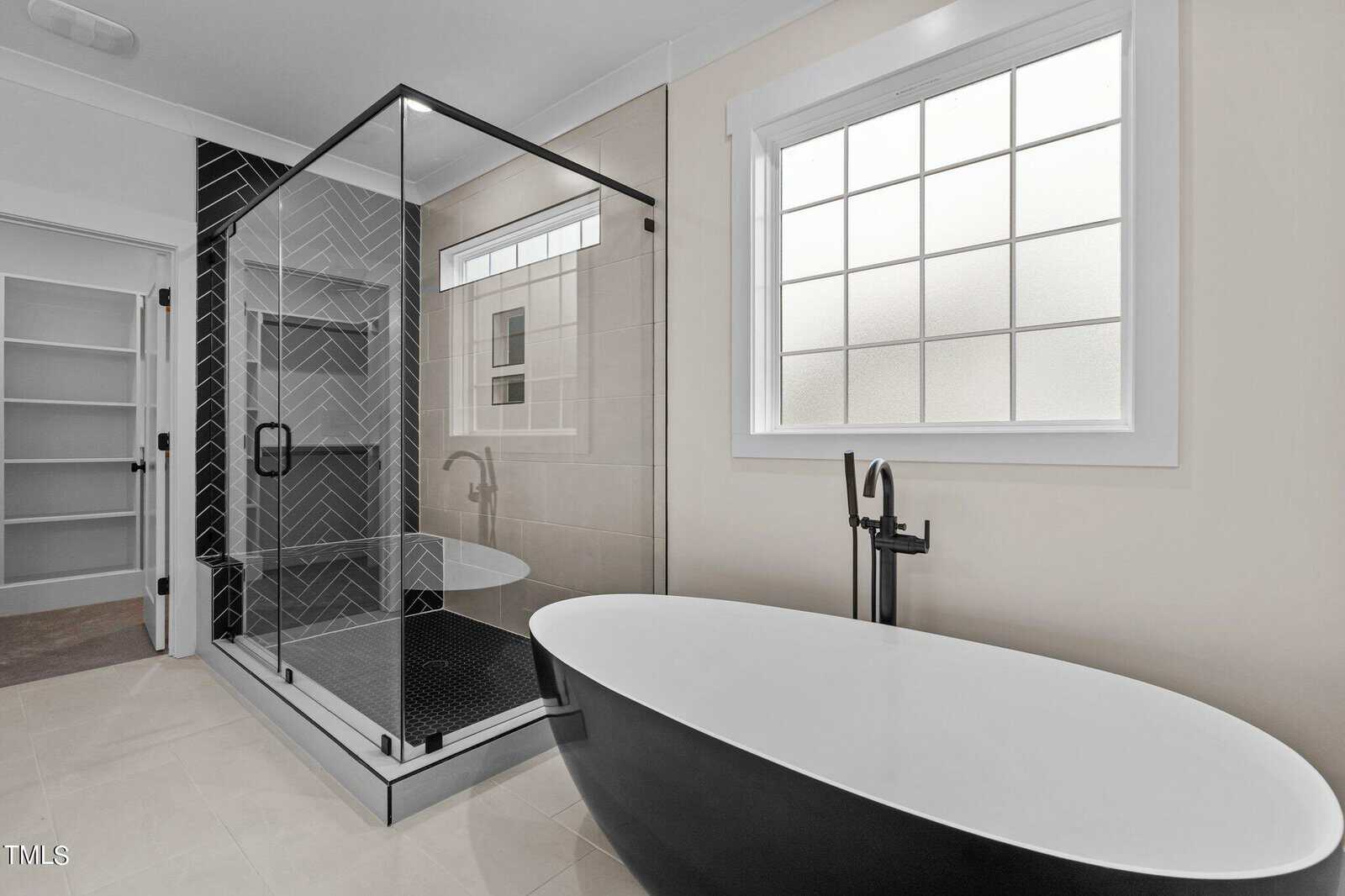 Modern bathroom with a black and white freestanding bathtub, a glass-enclosed shower with black herringbone tile, a large frosted window, and white shelves visible through an open door.