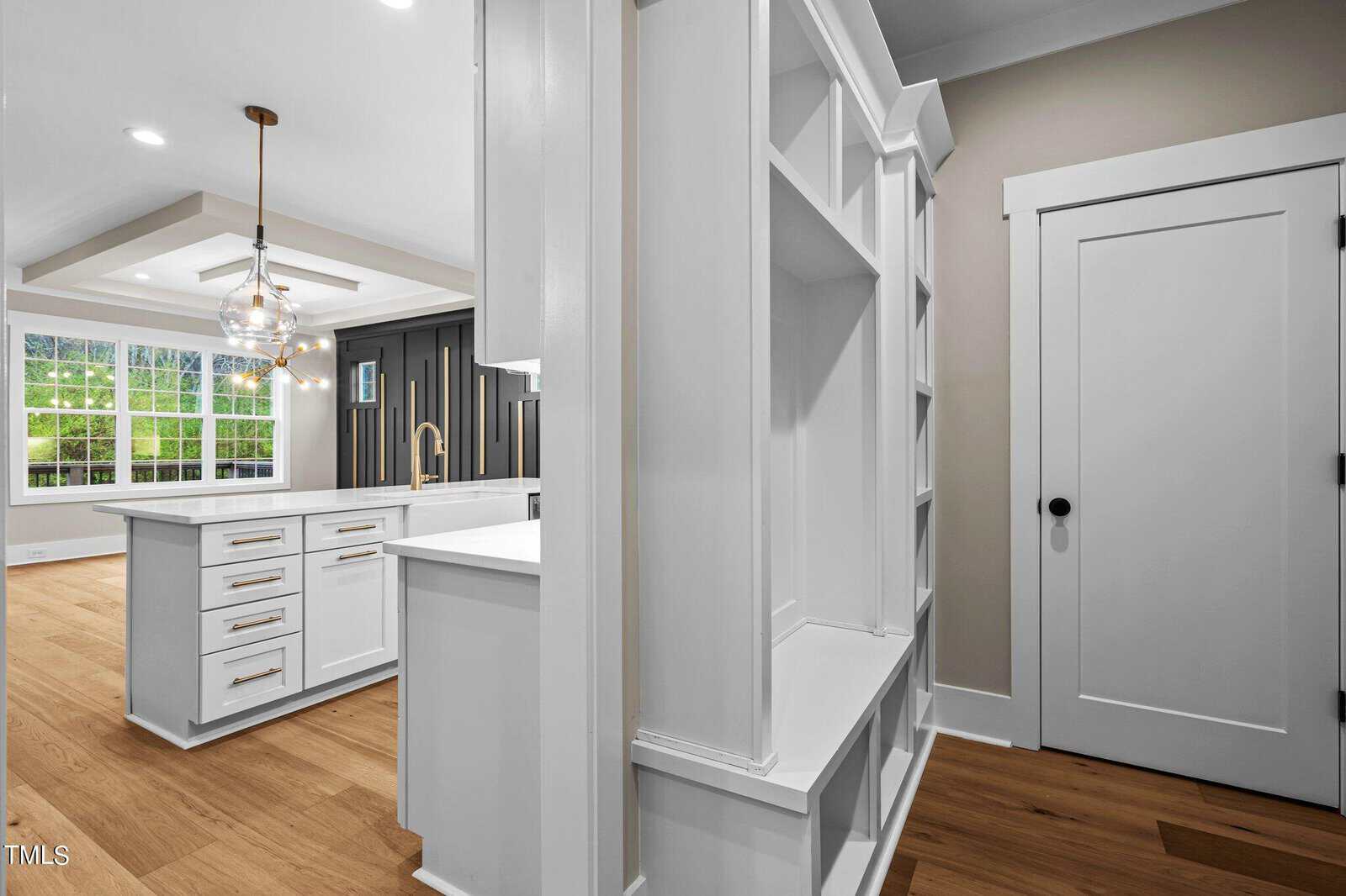 Bright, modern kitchen with white cabinetry, gold hardware, and a large island. Open shelving, light wood floors, and a geometric chandelier. Adjacent hallway with built-in shelves and a white door. Large windows let in natural light.