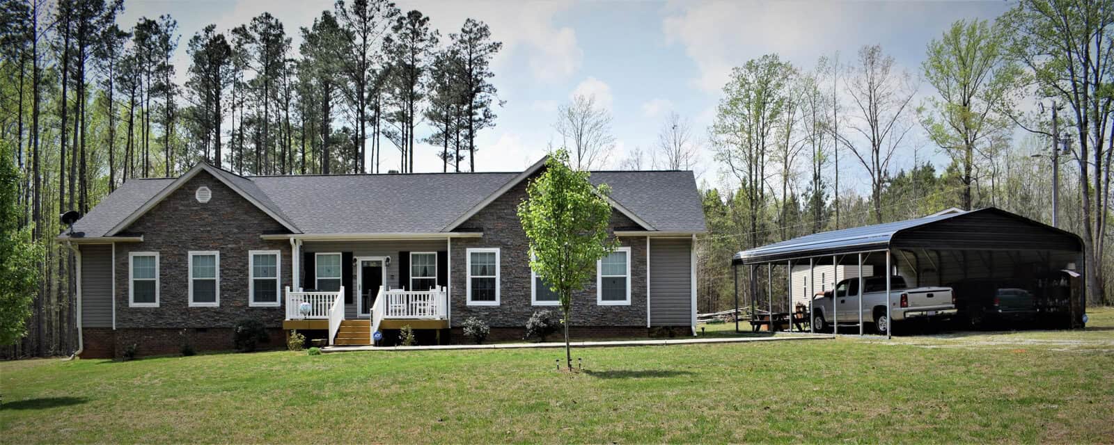 A single-story house with a brick frontage and tan siding stands in a clearing surrounded by tall trees. There is a small covered porch with white railings and a nearby carport sheltering a pickup truck alongside another vehicle. The yard is spacious and grassy.