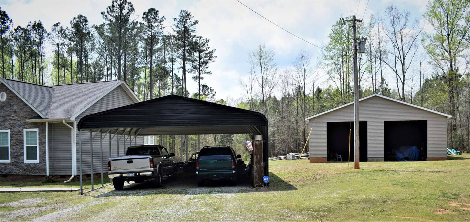 A house with gray siding and a stone accent wall is on the left. Two vehicles, a white truck and a green SUV, are parked under a black carport in the center. A separate garage building with two open bays is on the right, surrounded by a grassy yard and trees in the background.