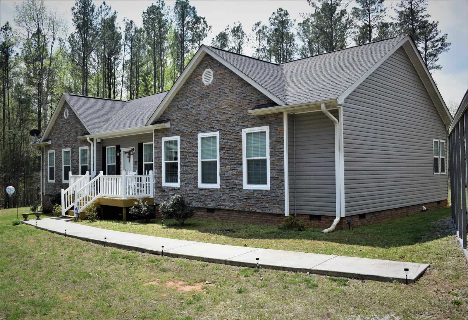 A single-story house with gray siding and stone accents. It has a small white front porch with steps and a ramp, surrounded by a grassy yard. Tall trees form a wooded backdrop. The house features four windows on the front side.